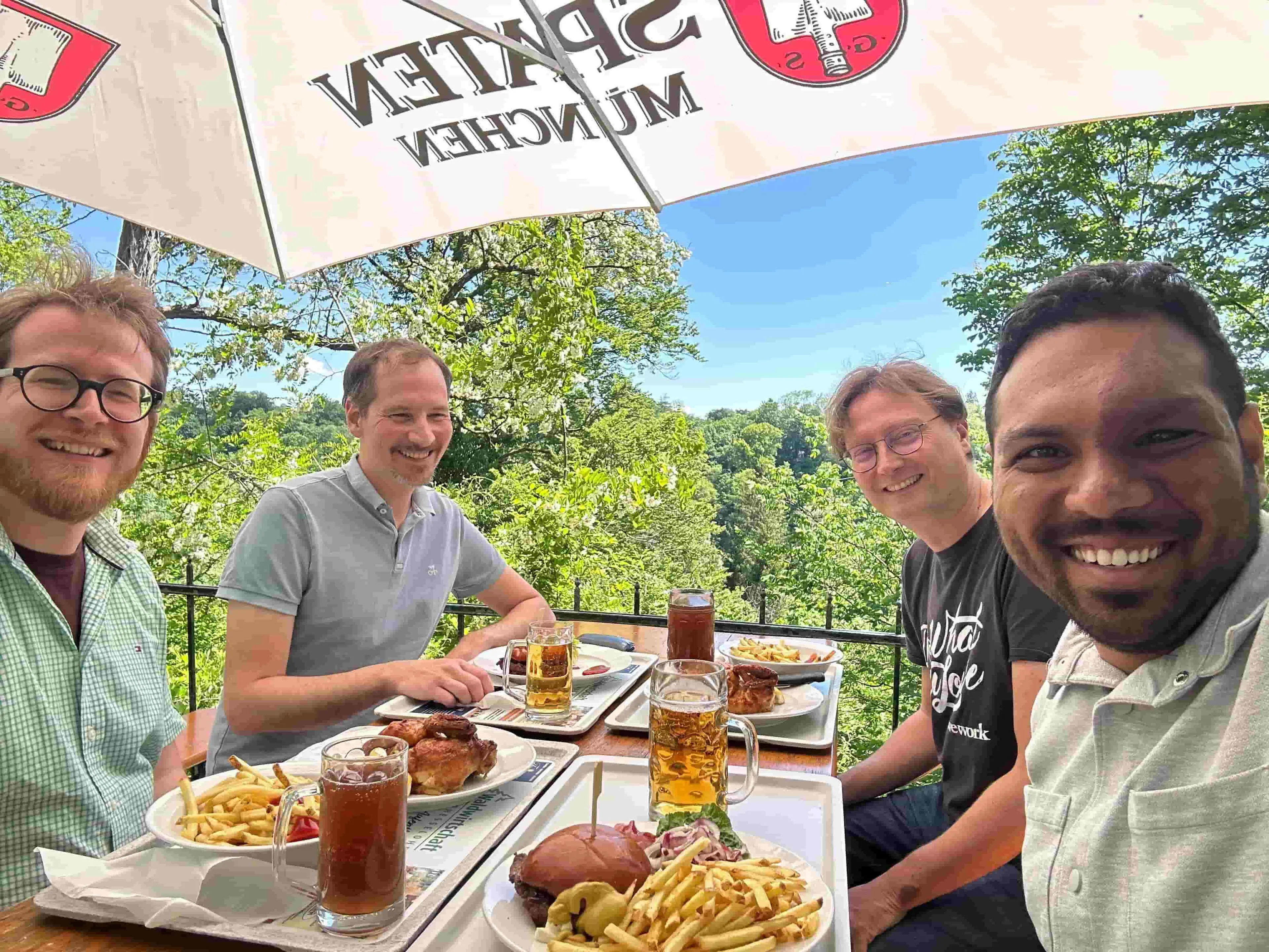 Four people having a meal, Manuel Krug, Arno Fleck, Thomas Schiwietz, and Anderson Vankayala enjoy burgers, fries, and classic German apfelschorle (apple juice and mineral water) 