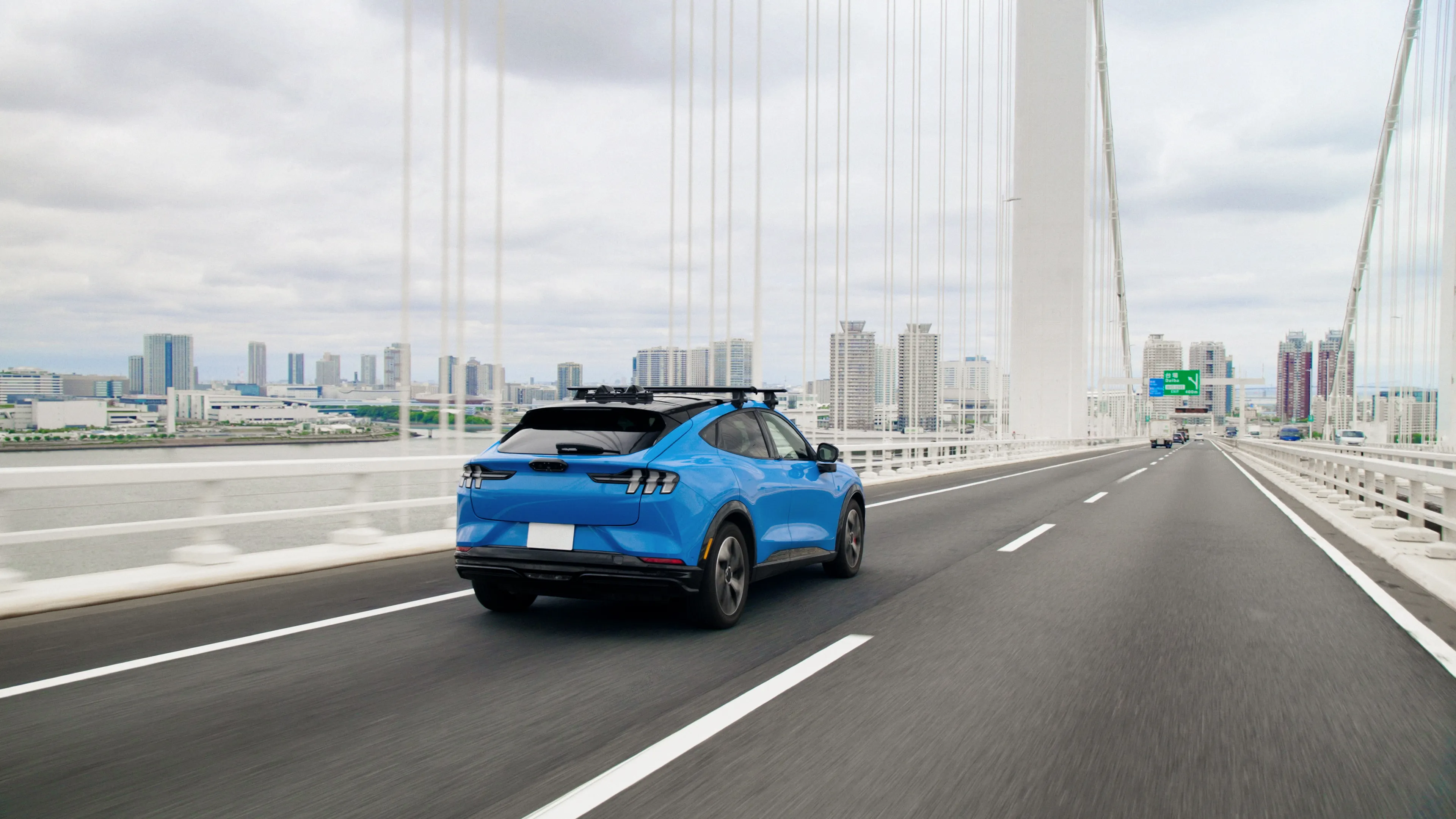Rear view of a blue SUV with a roof-mounted rack traveling along a wide suspension bridge, with a city skyline and overcast sky visible in the distance.