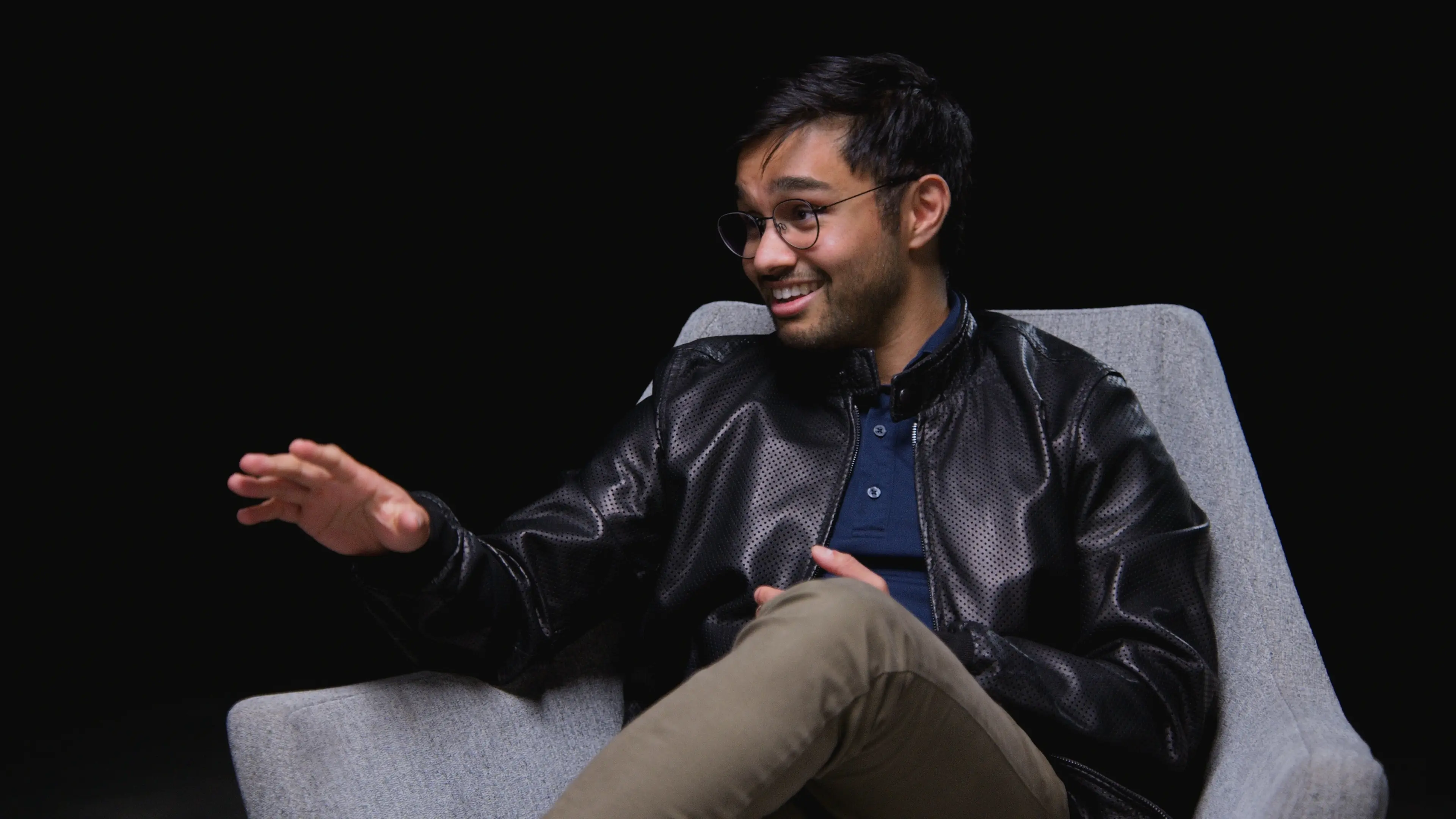 Malhar Patel smiles and gestures with one hand while seated in a gray chair against a black background.