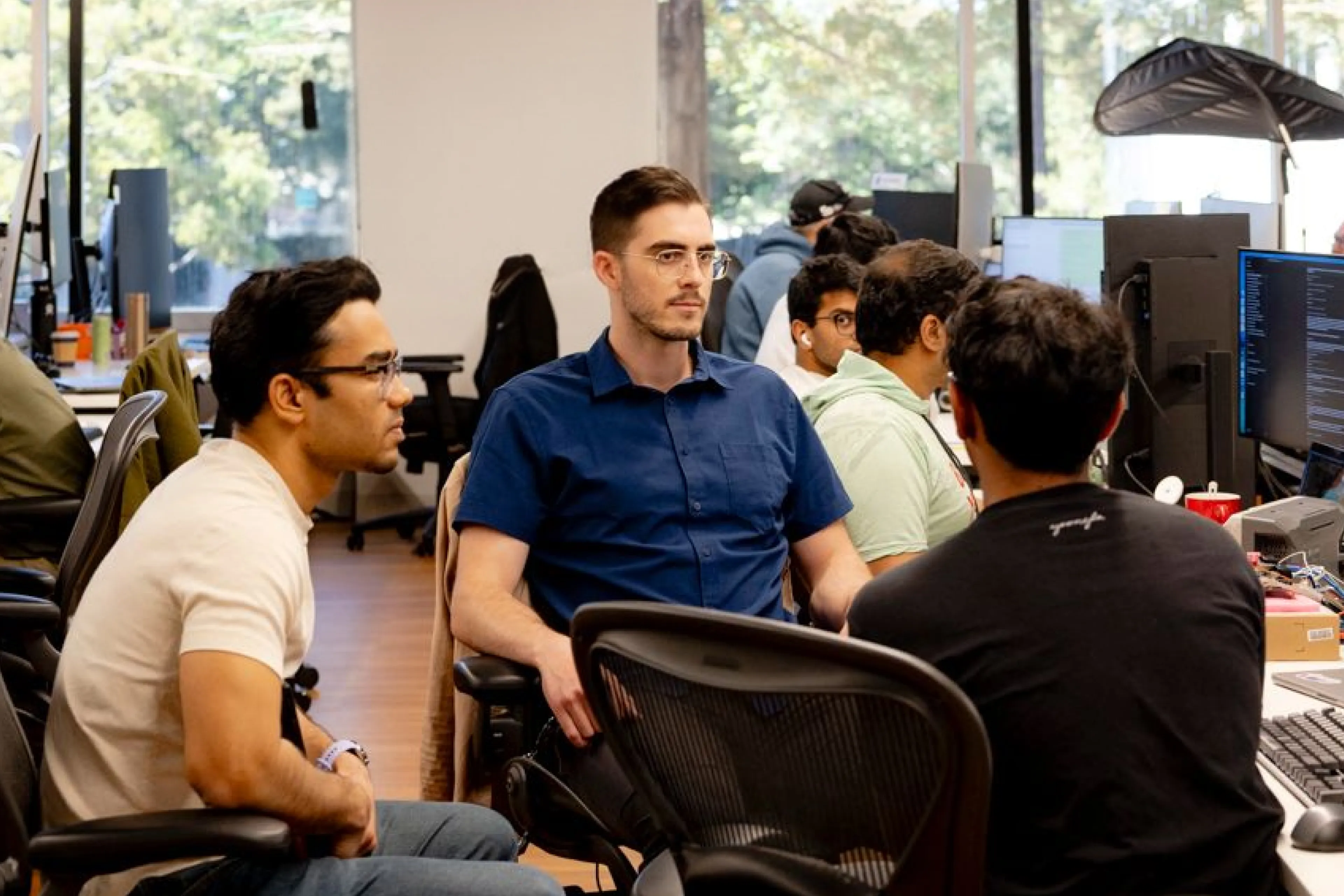 Coworkers talking at their desks in a tech office, with monitors and coding screens visible