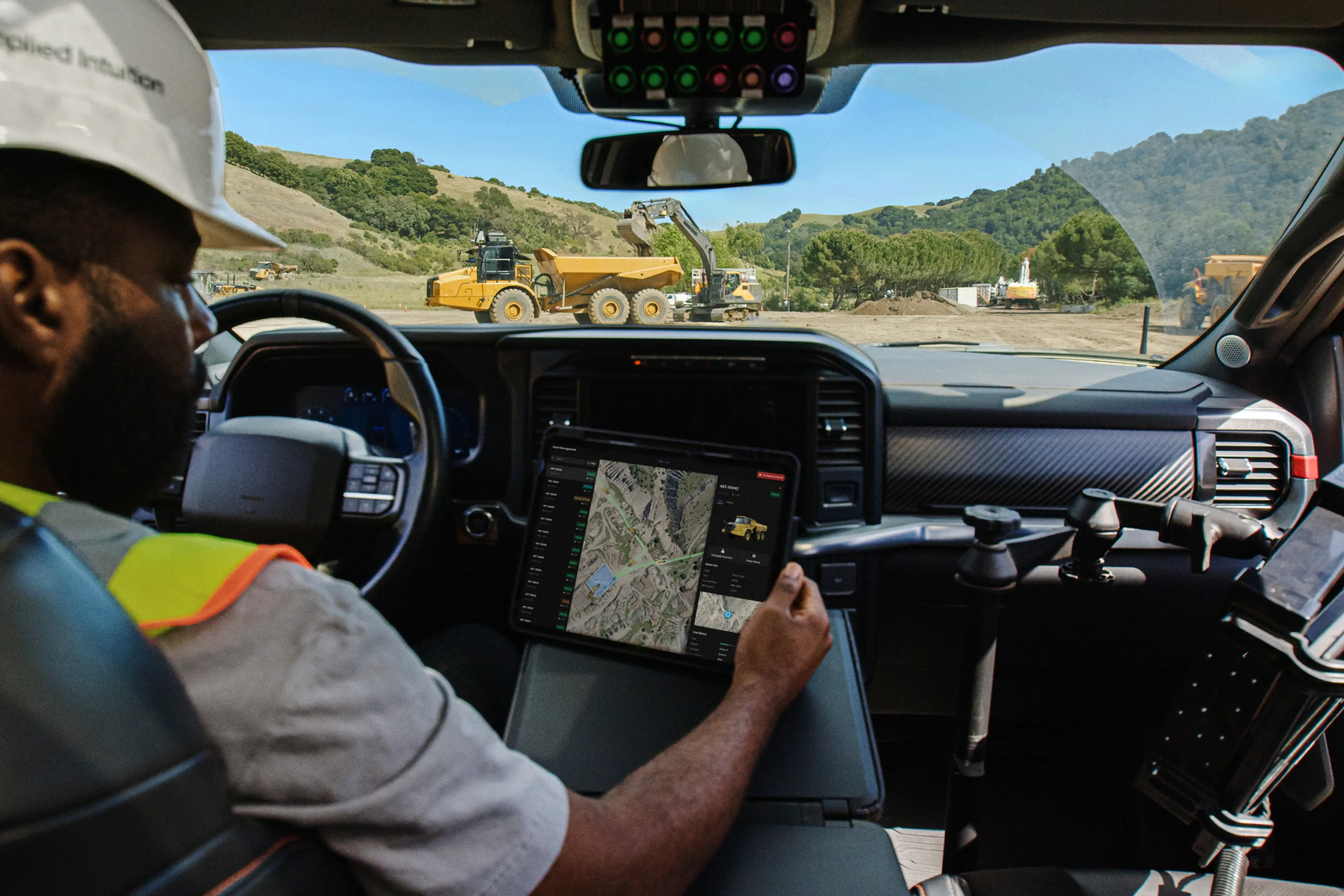 A construction worker sitting in a vehicle cab reviews a tablet showing a fleet management map, with heavy construction equipment visible through the windshield