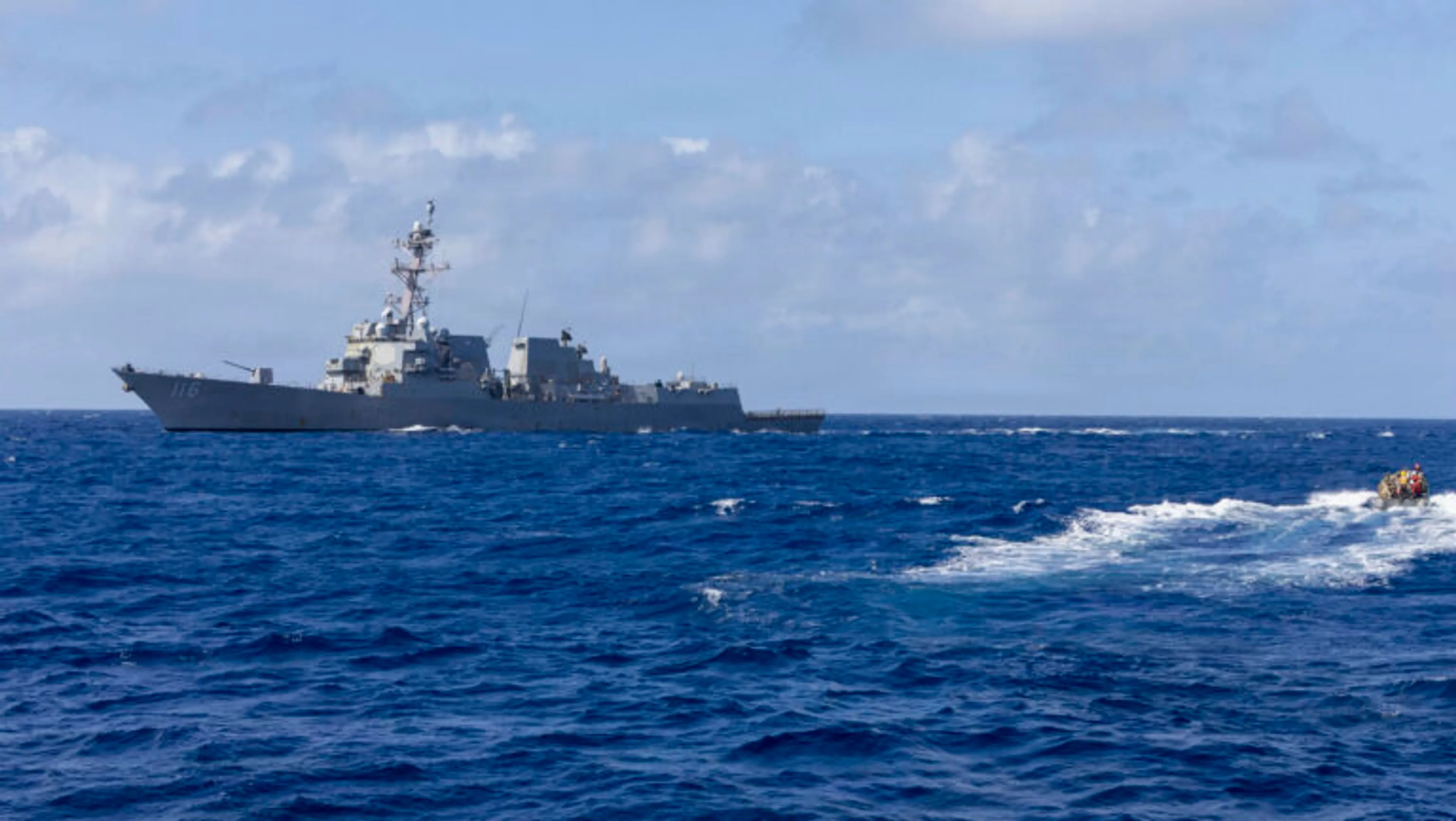 A U.S. Navy guided-missile destroyer (hull 116) underway on the open ocean, with a crewed rigid-hull inflatable boat speeding across the water in the foreground.