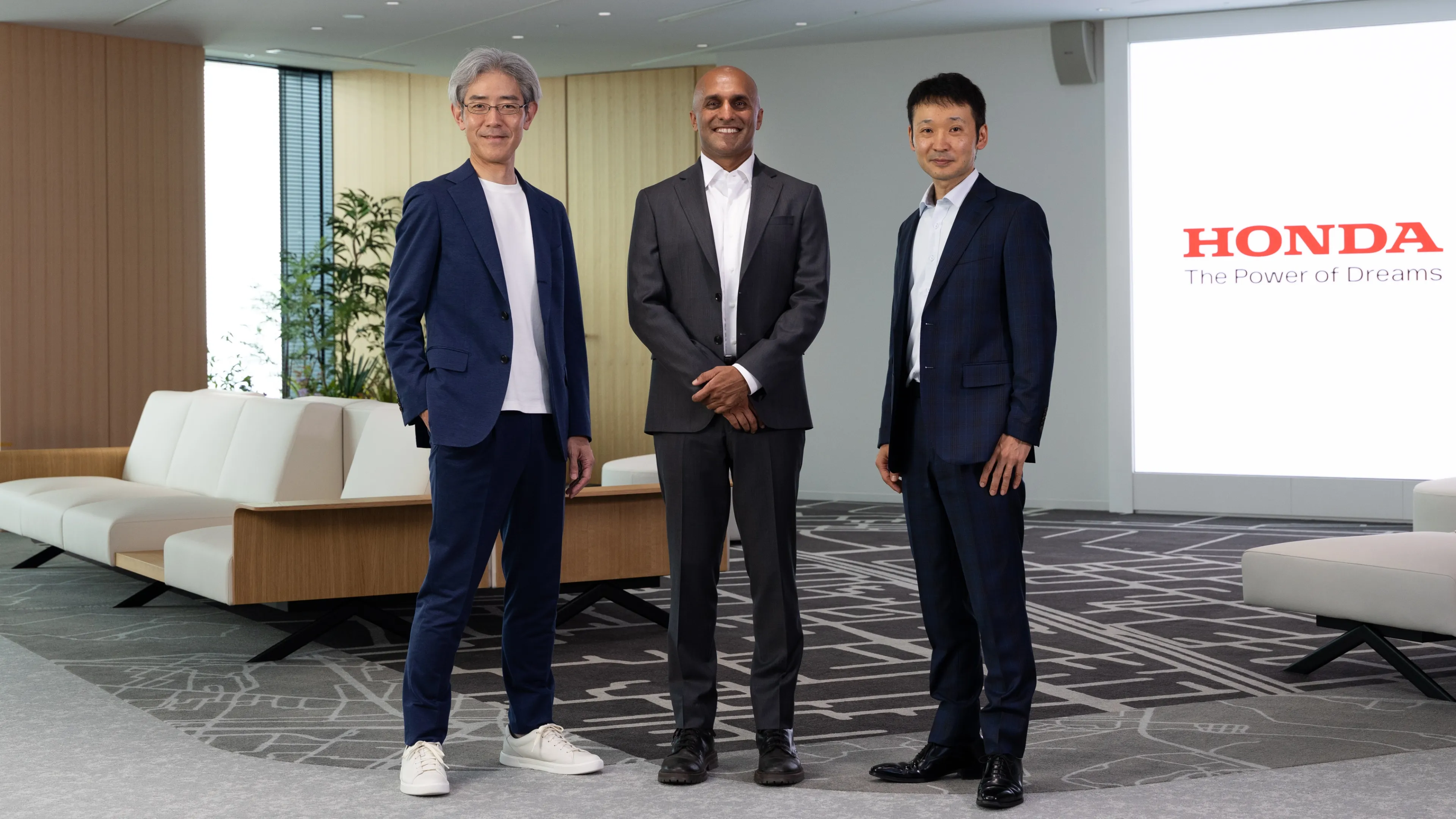 Shinichi Arie of Honda R&D, Qasar Younis of Applied Intuition, and Mahito Shikama of Honda, left to right, in an office meeting space with a Honda logo displayed on a screen behind them.