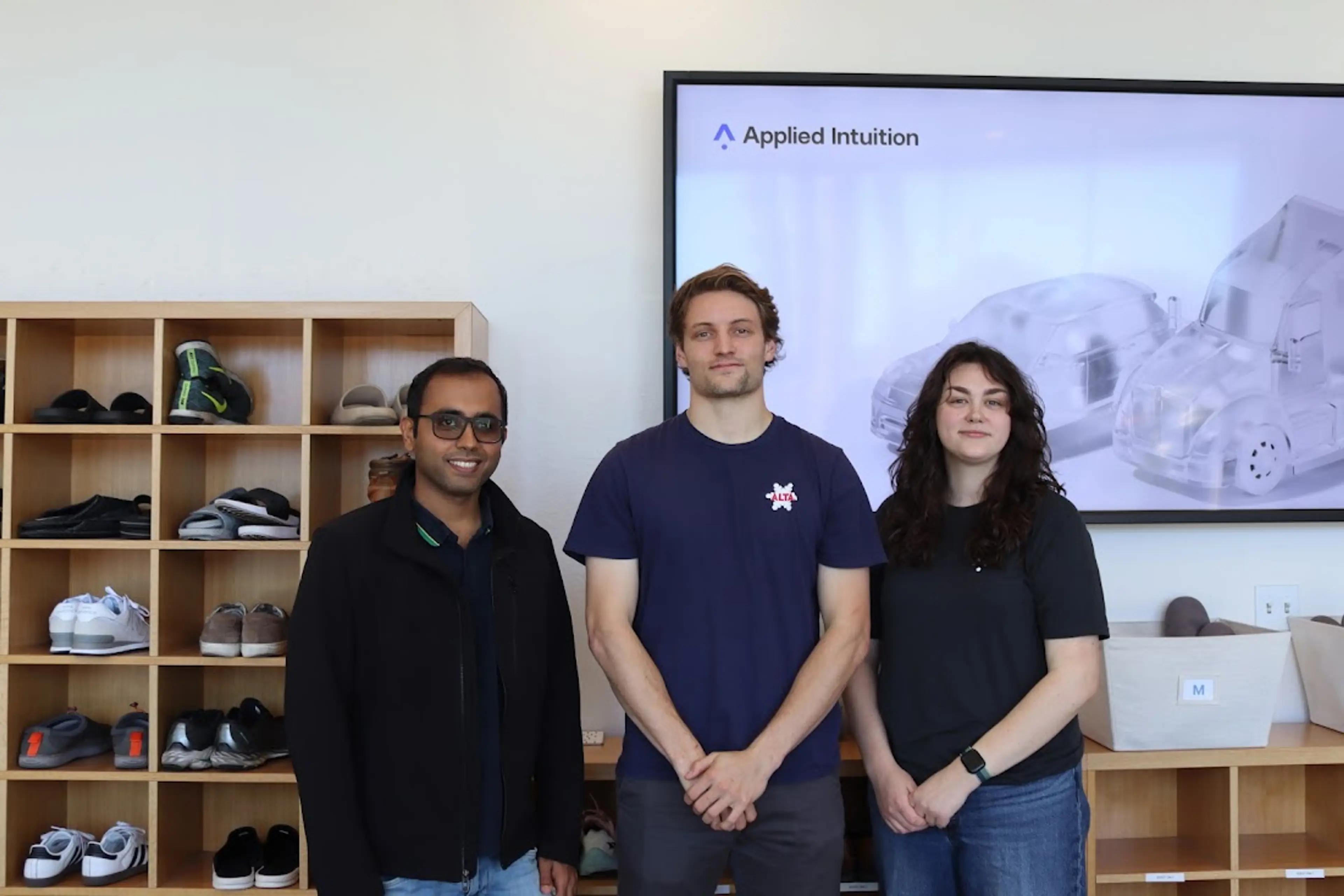 Three people stand facing the camera in an office lobby, in front of cubbies holding shoes