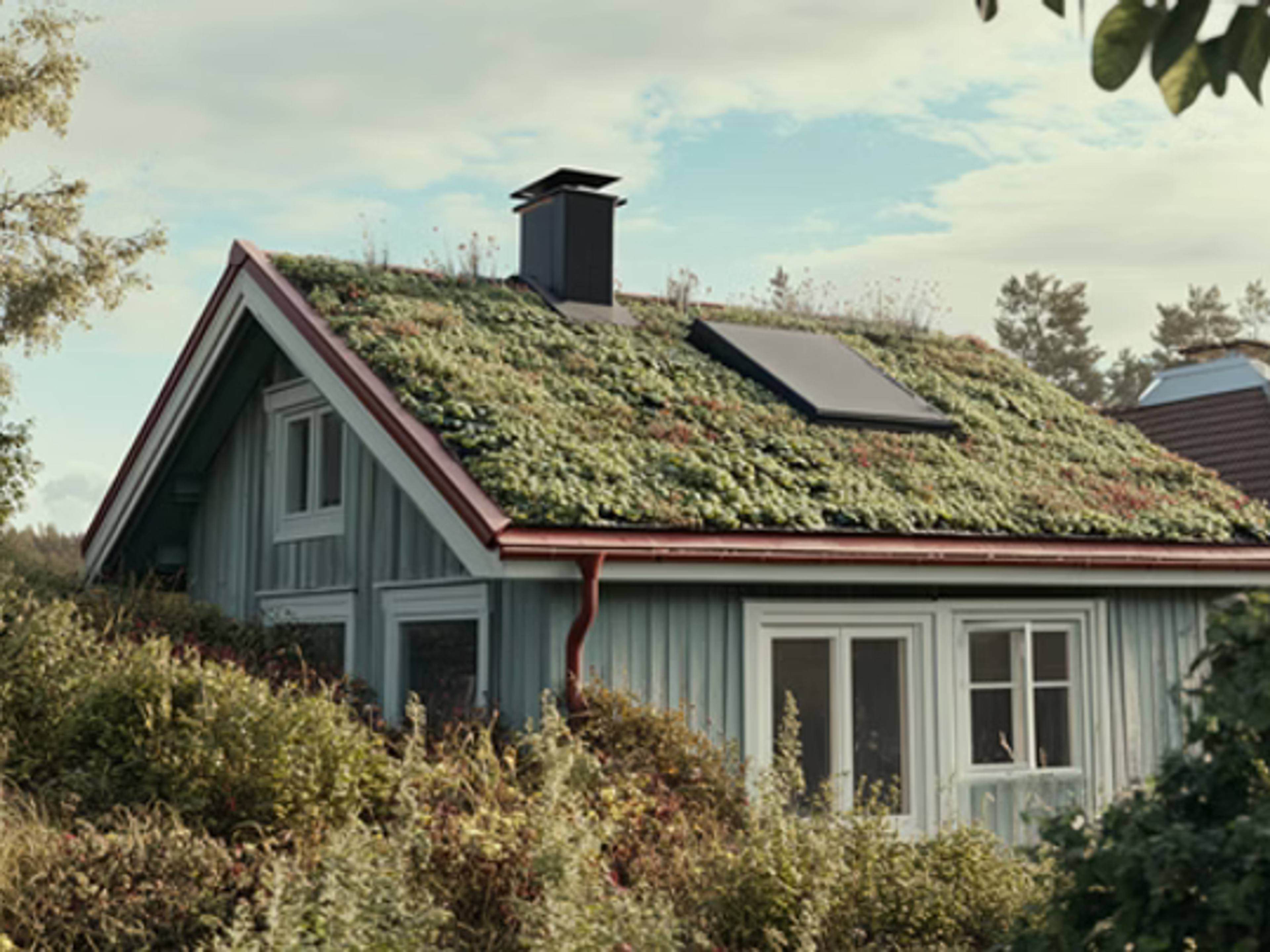 Grått hus med grönt sedumtak, skorsten och solpanel, omgivet av grönska