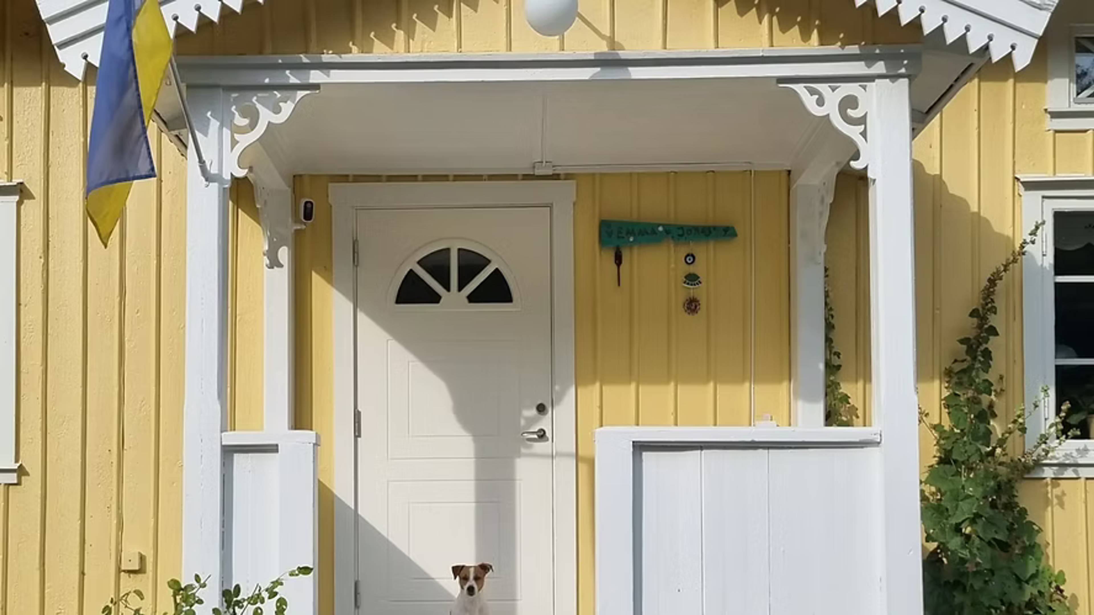 Gult trähus med vit farstukvist, dekorativa snickerier och en hund vid dörren