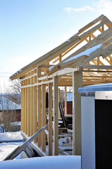 Timmerstomme under uppbyggnad täckt av snö, med rött hus i bakgrunden