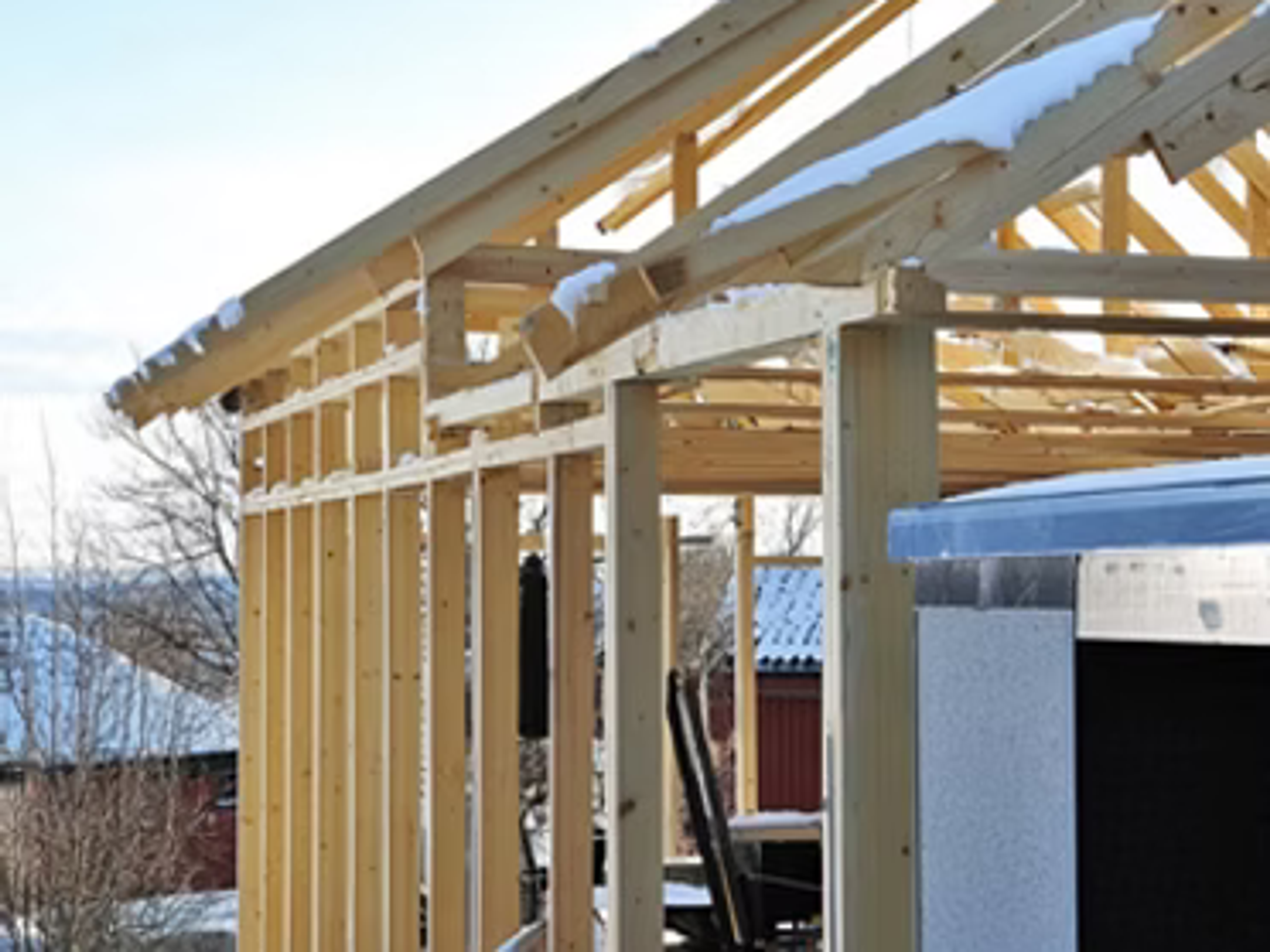 Timmerstomme under uppbyggnad täckt av snö, med rött hus i bakgrunden