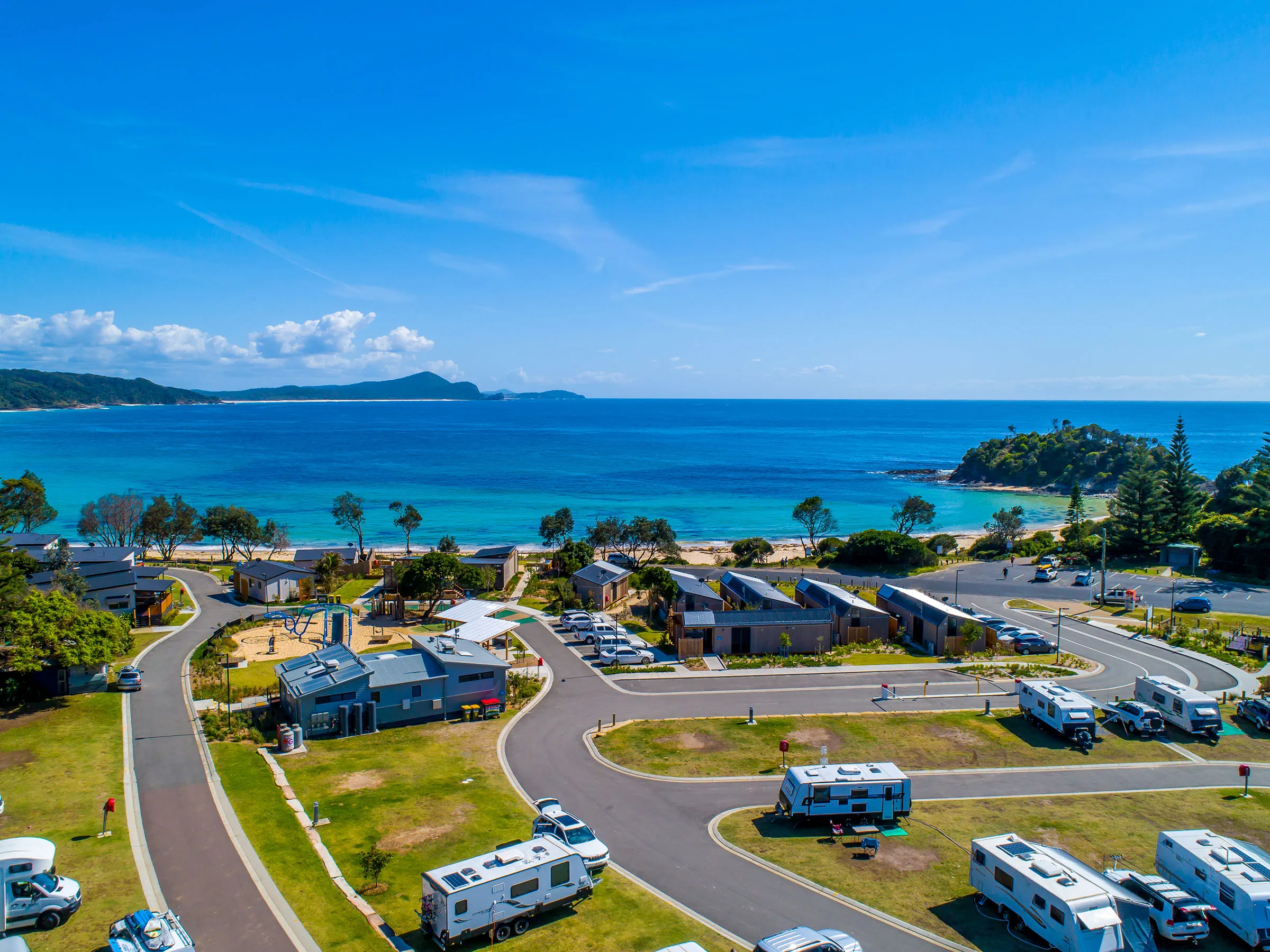A scenic view of Reflections Seal Rocks, showcasing a picturesque coastal camping area with caravans, cabins, and a stunning backdrop of turquoise ocean and lush greenery.