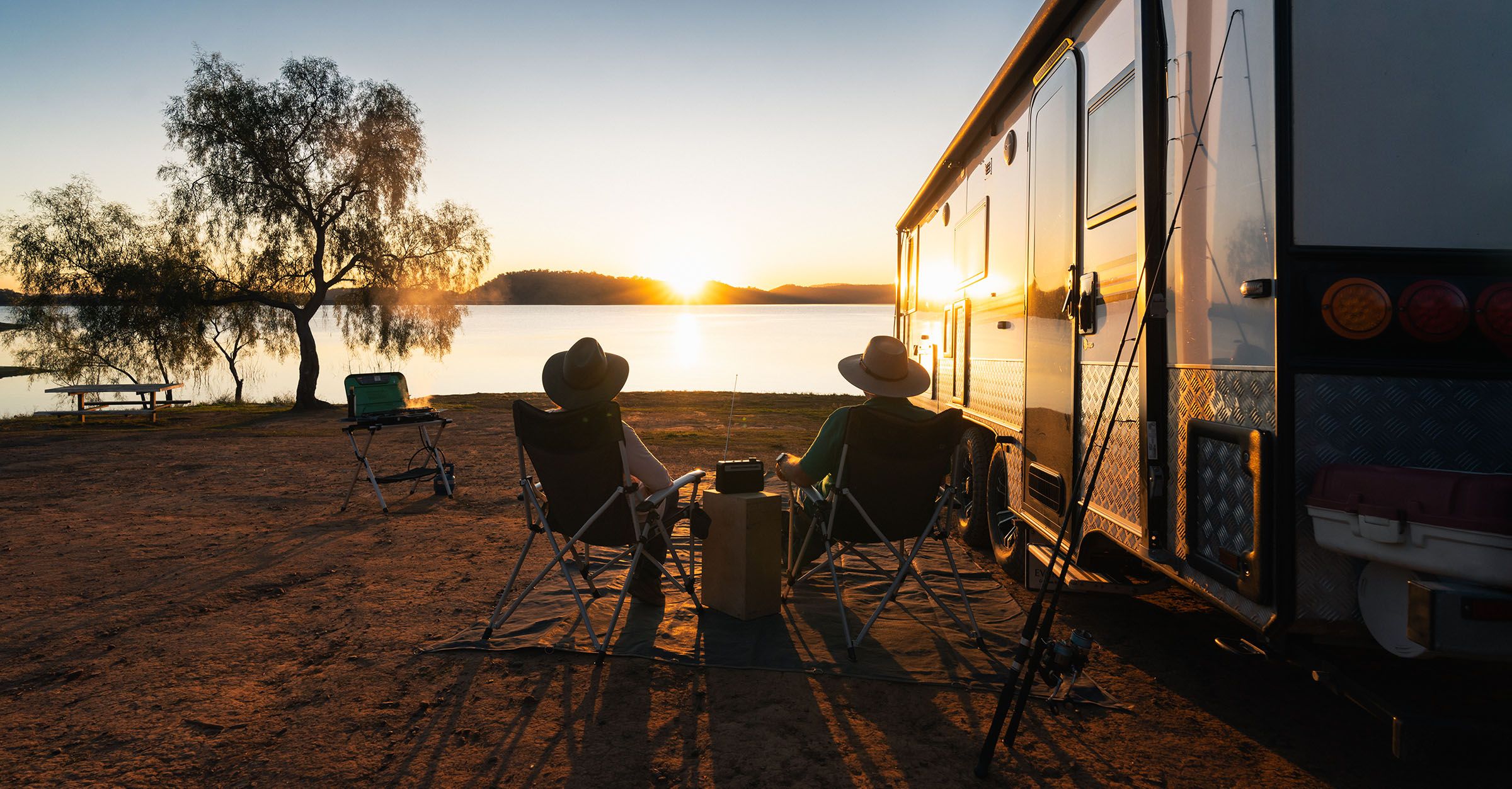 Two campers relax by their caravan at Reflections Holiday Lake Glenbawn, enjoying a tranquil lakeside sunset with a fishing rod nearby.