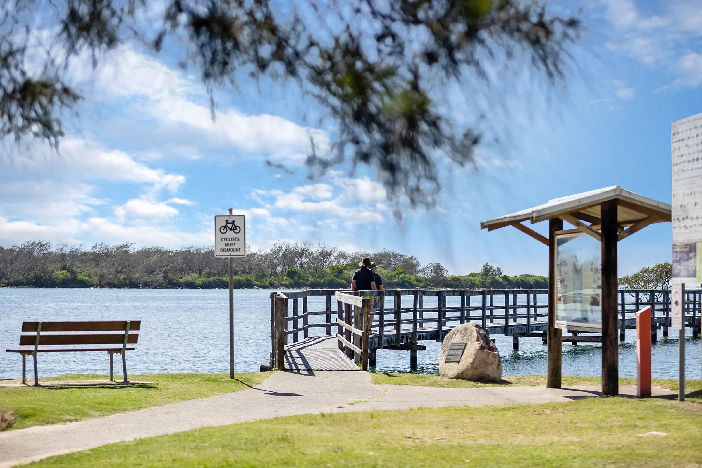 scenic waterfront boardwalk and walking path at Reflections Urunga Caravan Park, with signage and seating overlooking the river