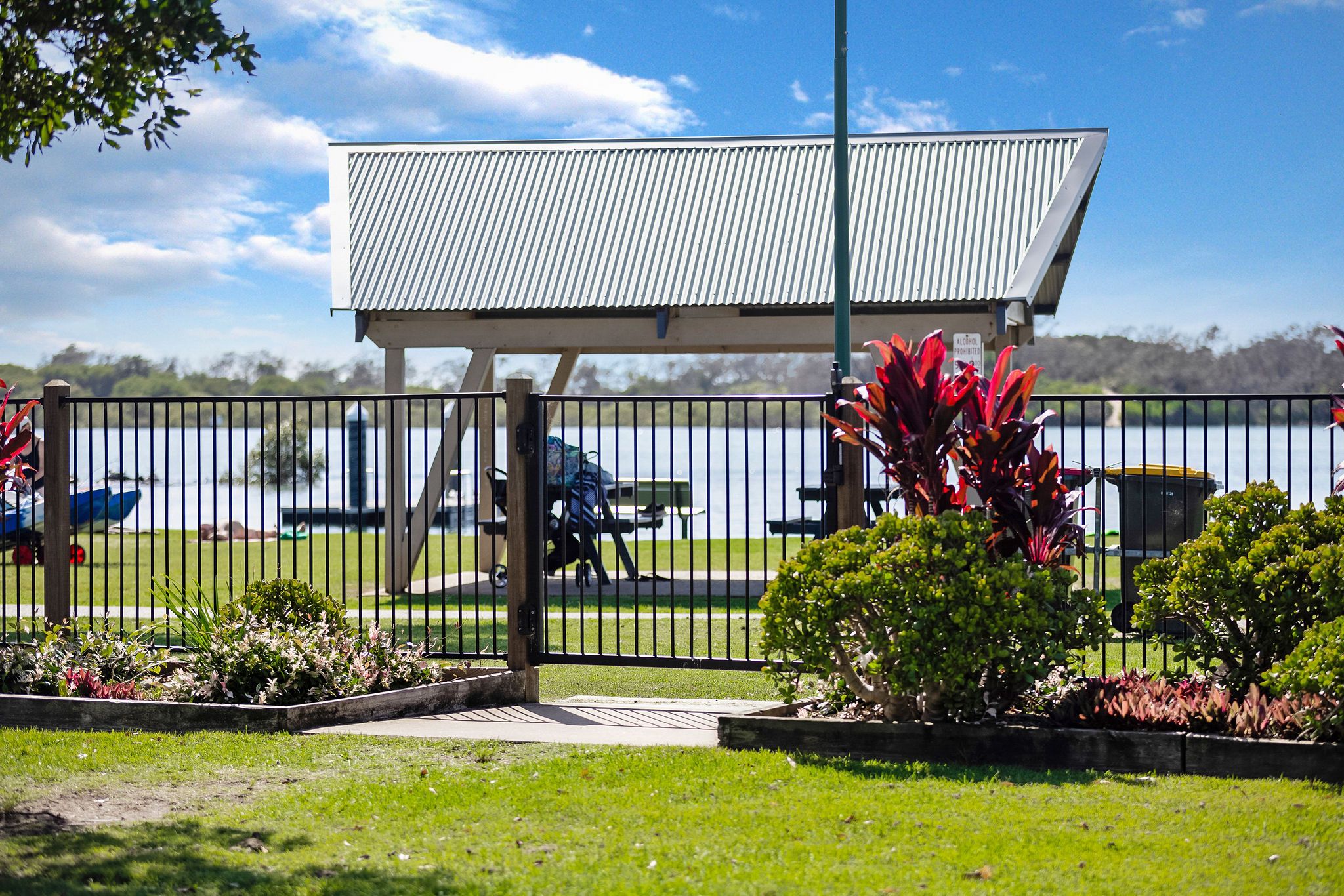 fenced picnic area by the river with a covered shelter and lush garden surroundings at Reflections Urunga Caravan Park