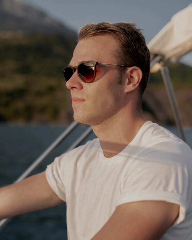 Homme en lunettes de soleil sur un voilier avec montagnes alpines en arrière-plan, lumière dorée — photographie de voyage par Paul Piccolini