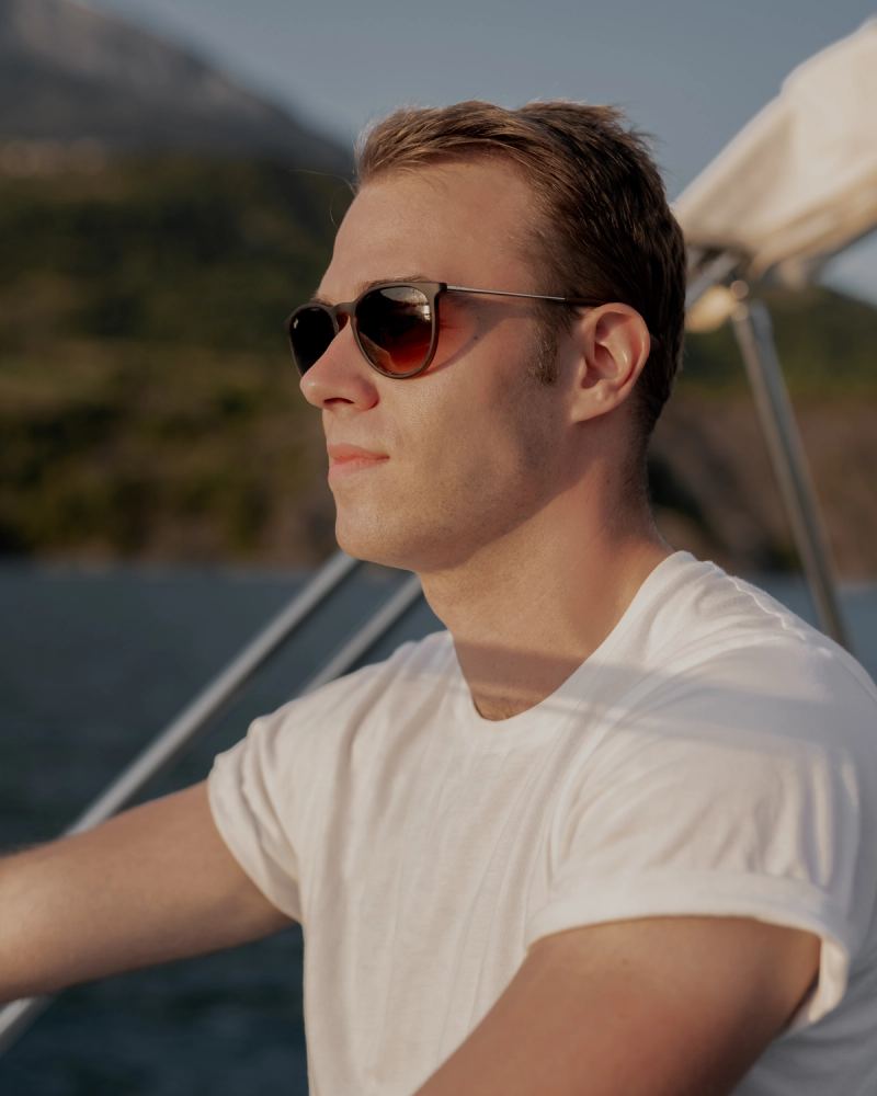 Homme en lunettes de soleil sur un voilier avec montagnes alpines en arrière-plan, lumière dorée — photographie de voyage par Paul Piccolini