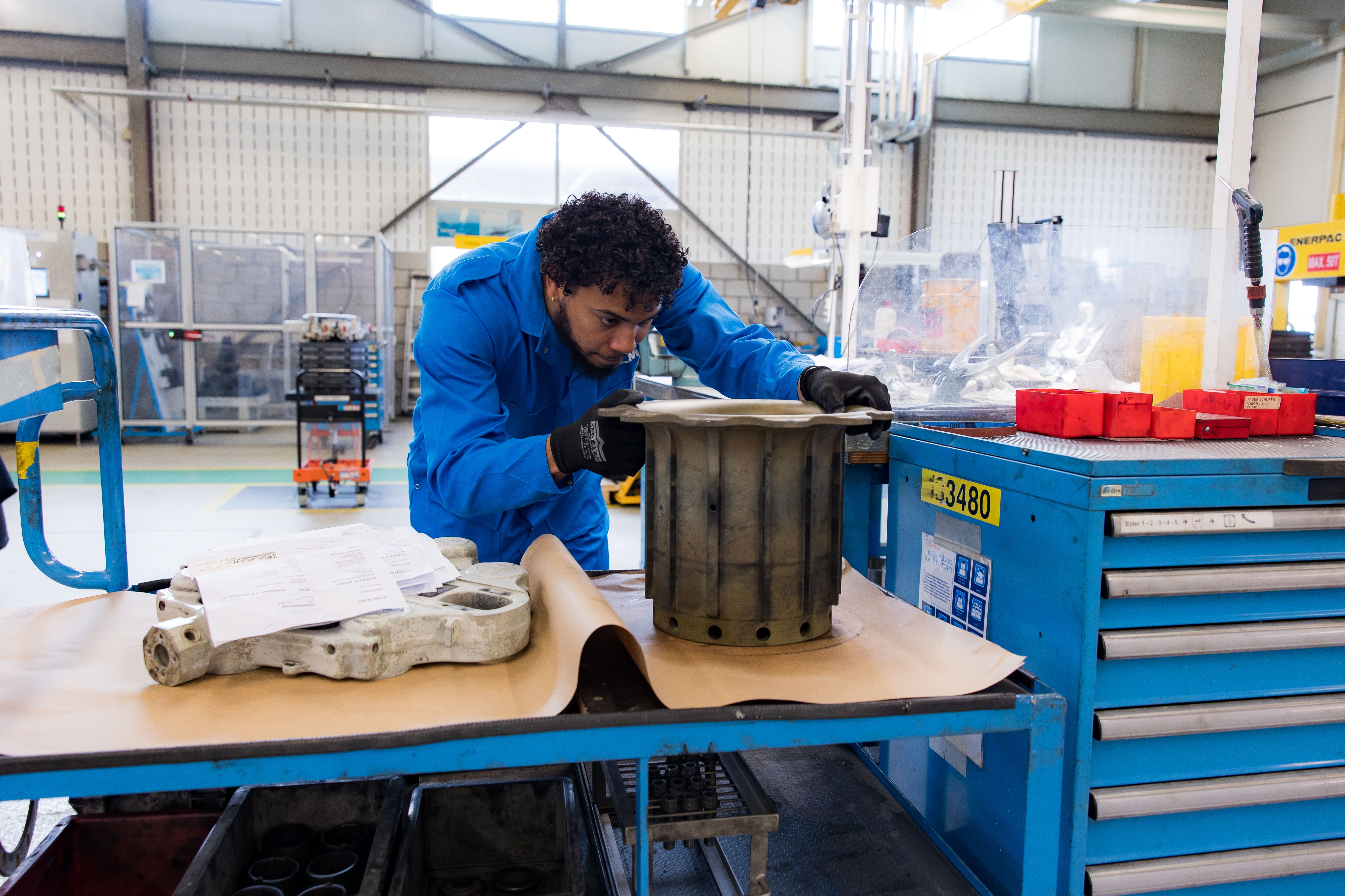 A mechanic in a blue overall is inspecting a large metal aircraft part on a workbench in a workshop
