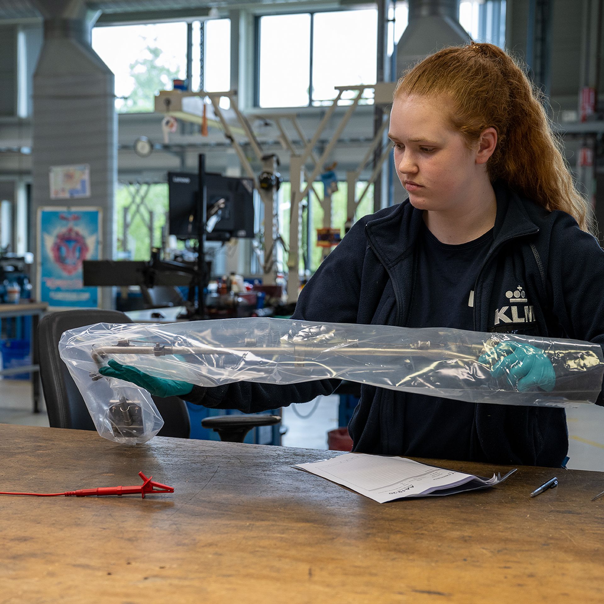 A woman in a KLM jacket is working at a workbench with a packaged aircraft part in a workshop.
