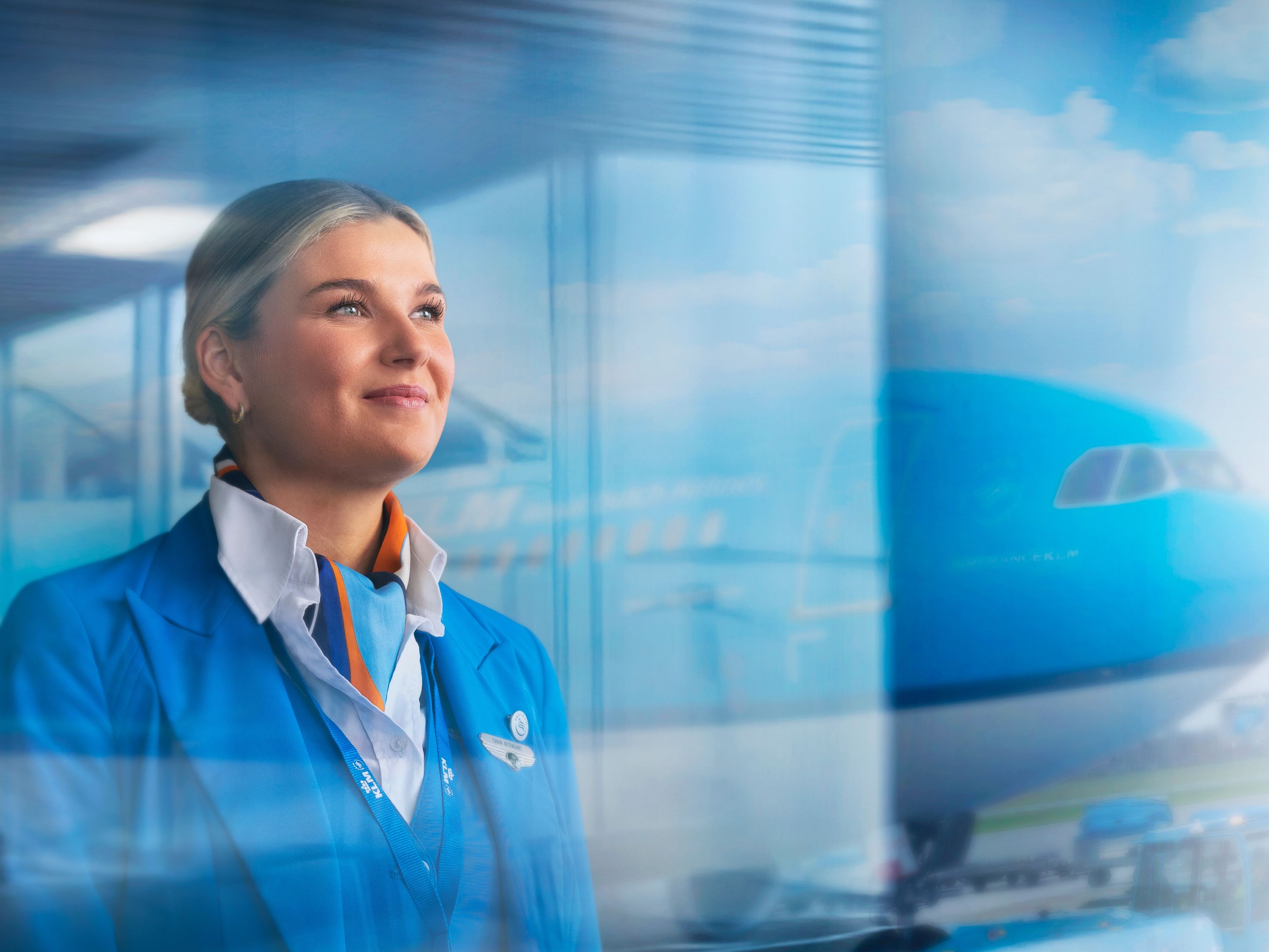 KLM cabin attendant die uit een raam kijkt met een vliegtuig op de achtergrond
