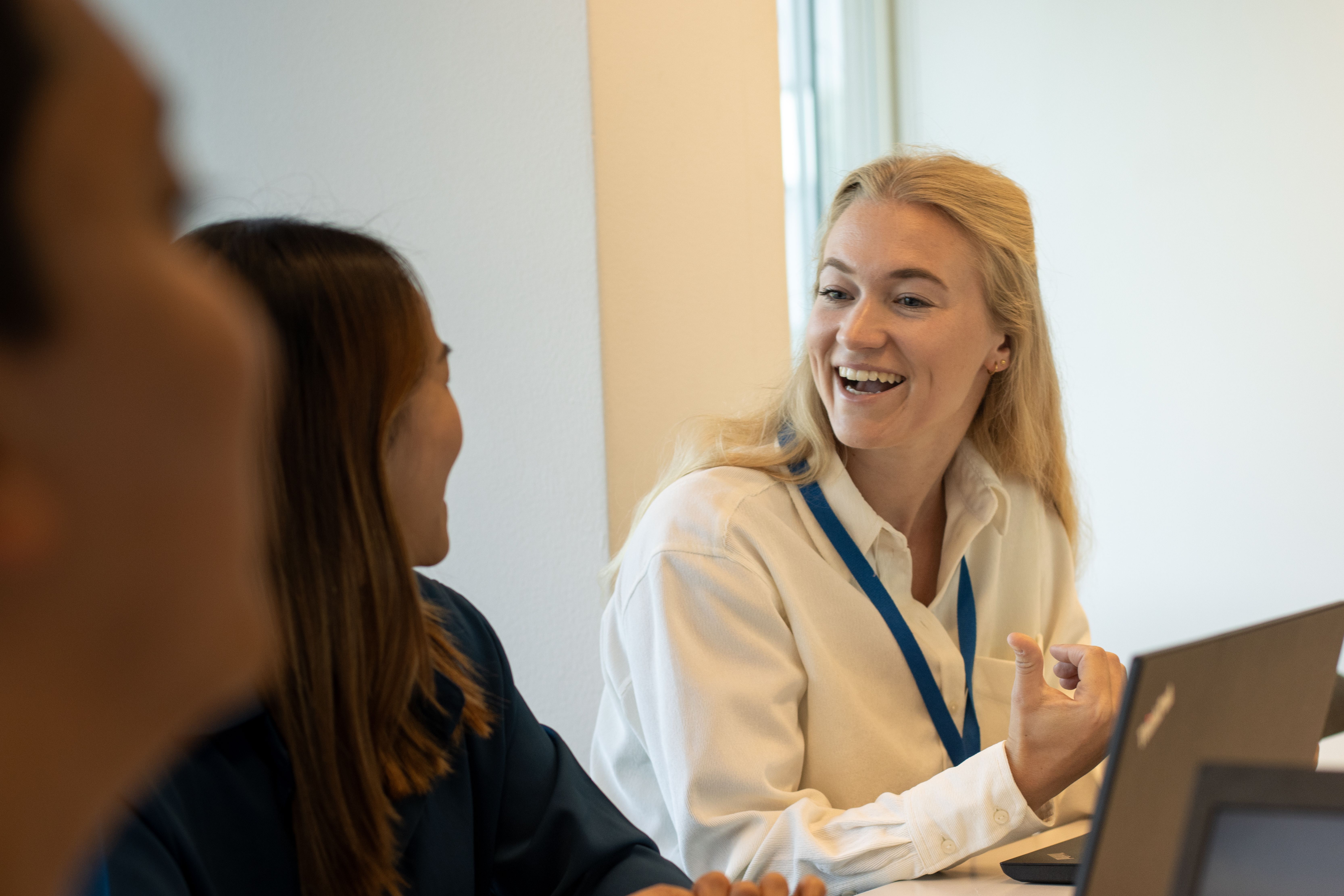 Een lachende blonde vrouw in een witte blouse met een blauw KLM-logo kijkt naar de collega die links van haar zit