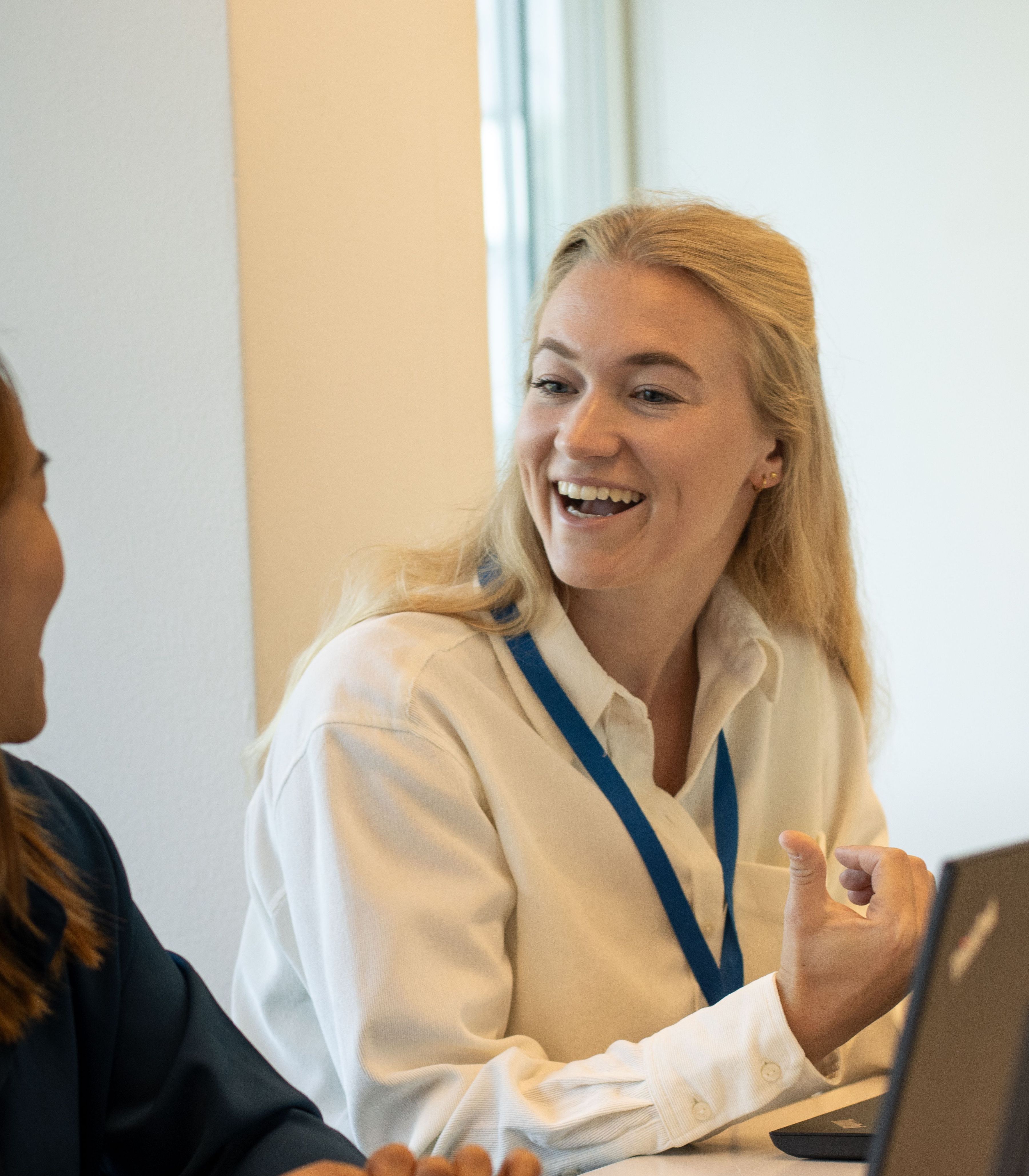 Een lachende blonde vrouw in een witte blouse met een blauw KLM-logo kijkt naar de collega die links van haar zit