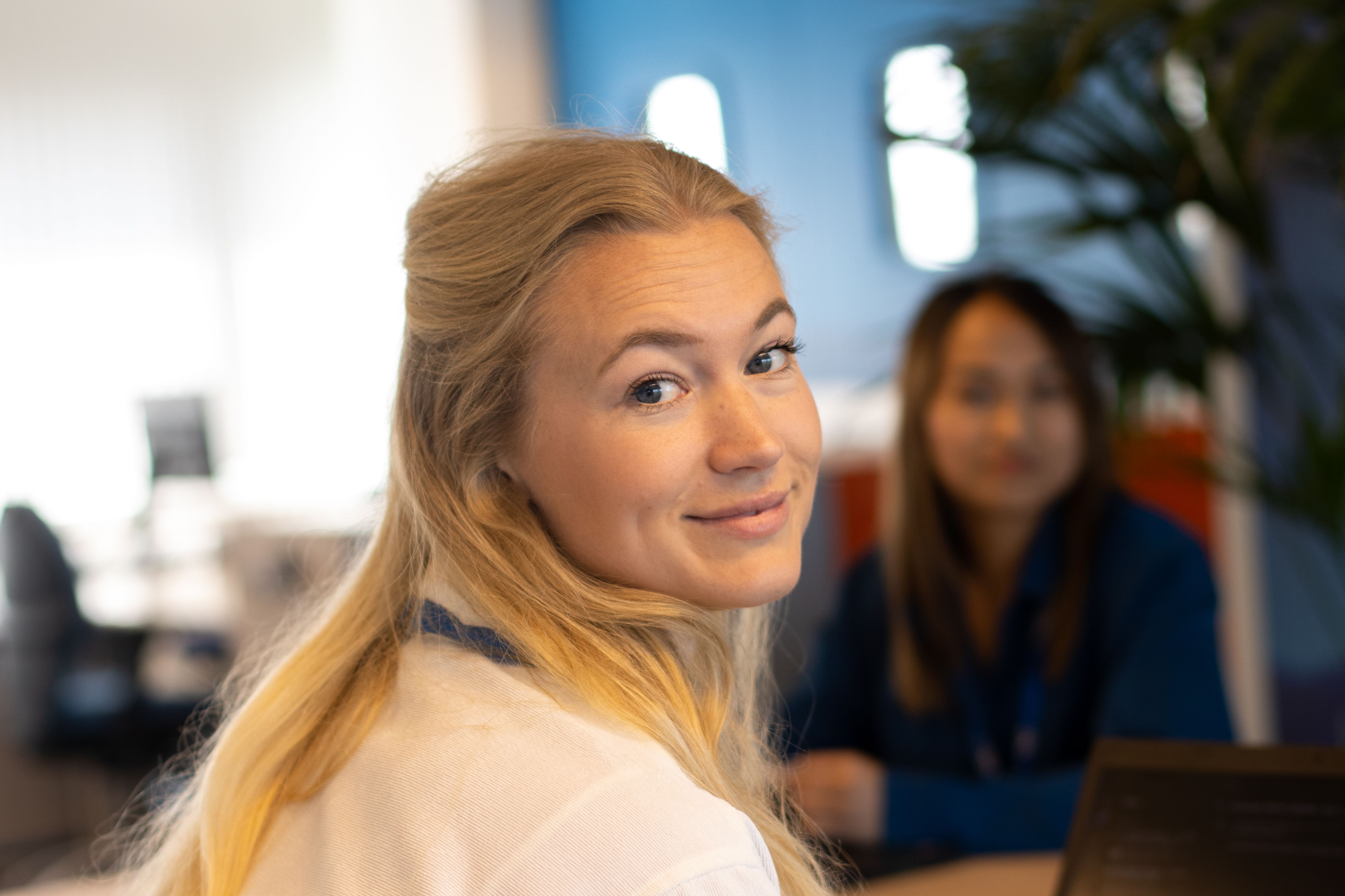 Lachende vrouw met lang blond haar die over haar schouder kijkt, met een andere vrouw vaag op de achtergrond.