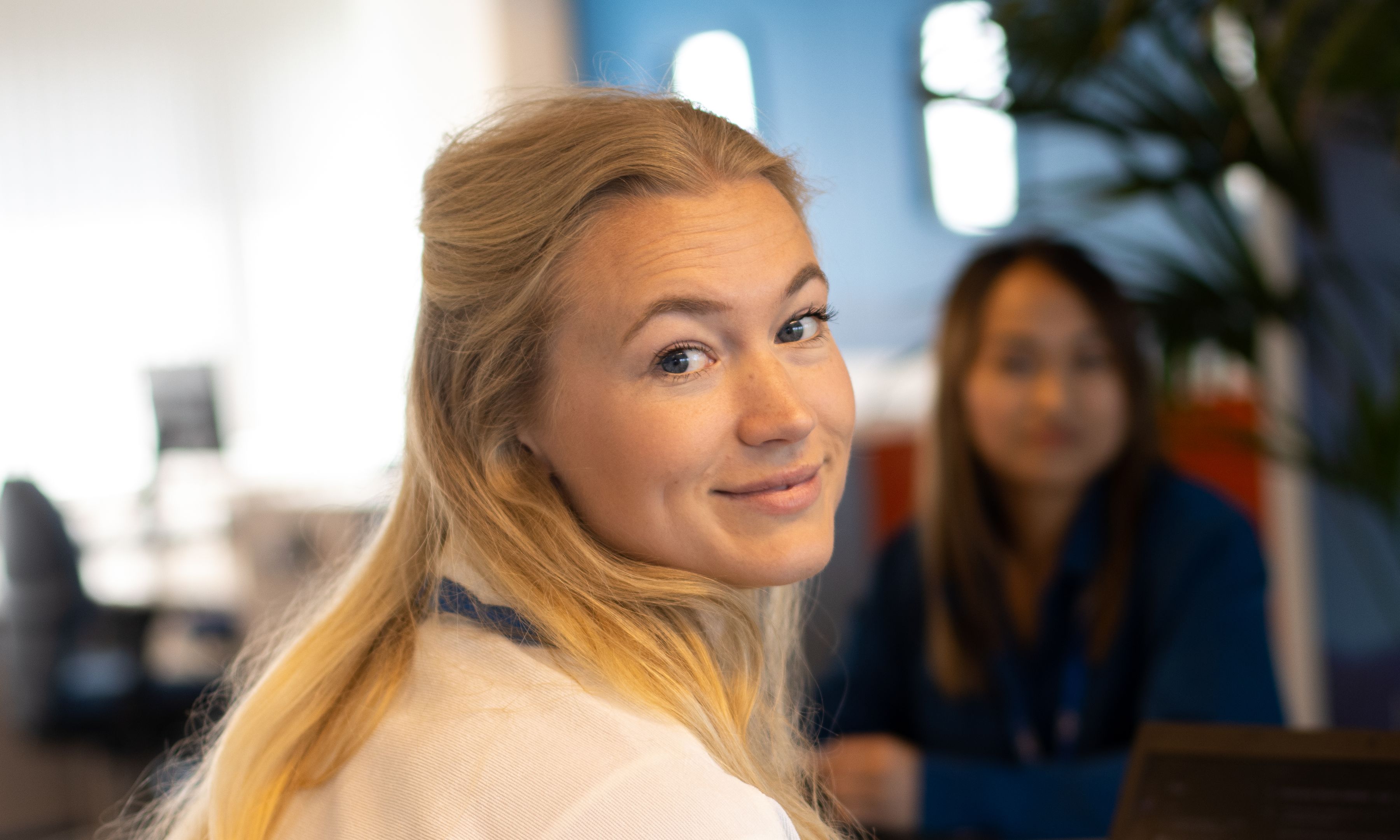 Lachende vrouw met lang blond haar die over haar schouder kijkt, met een andere vrouw vaag op de achtergrond.