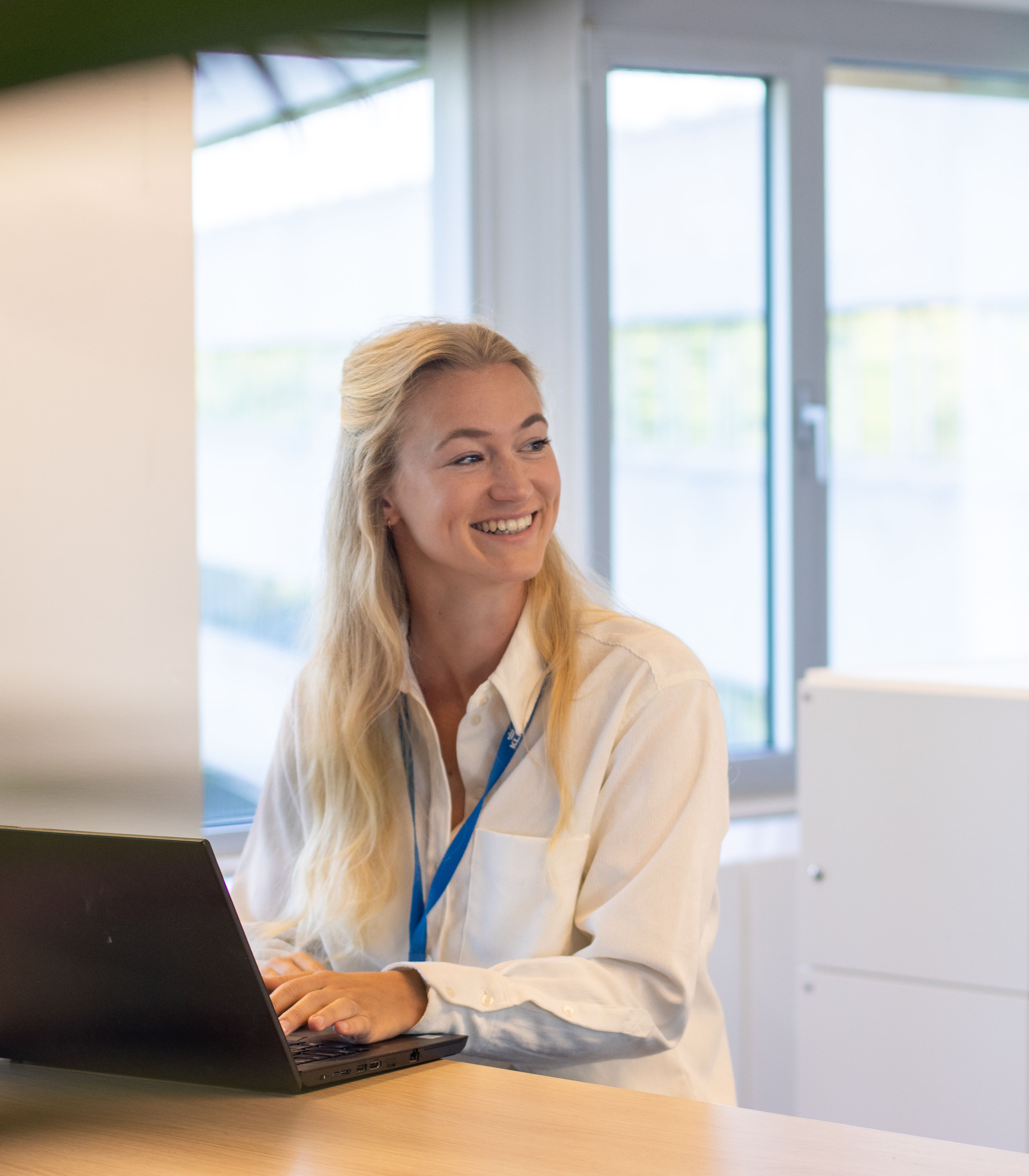 Woman smiling while working on a laptop in a bright office space, with a colleague holding a large paper roll in the background.