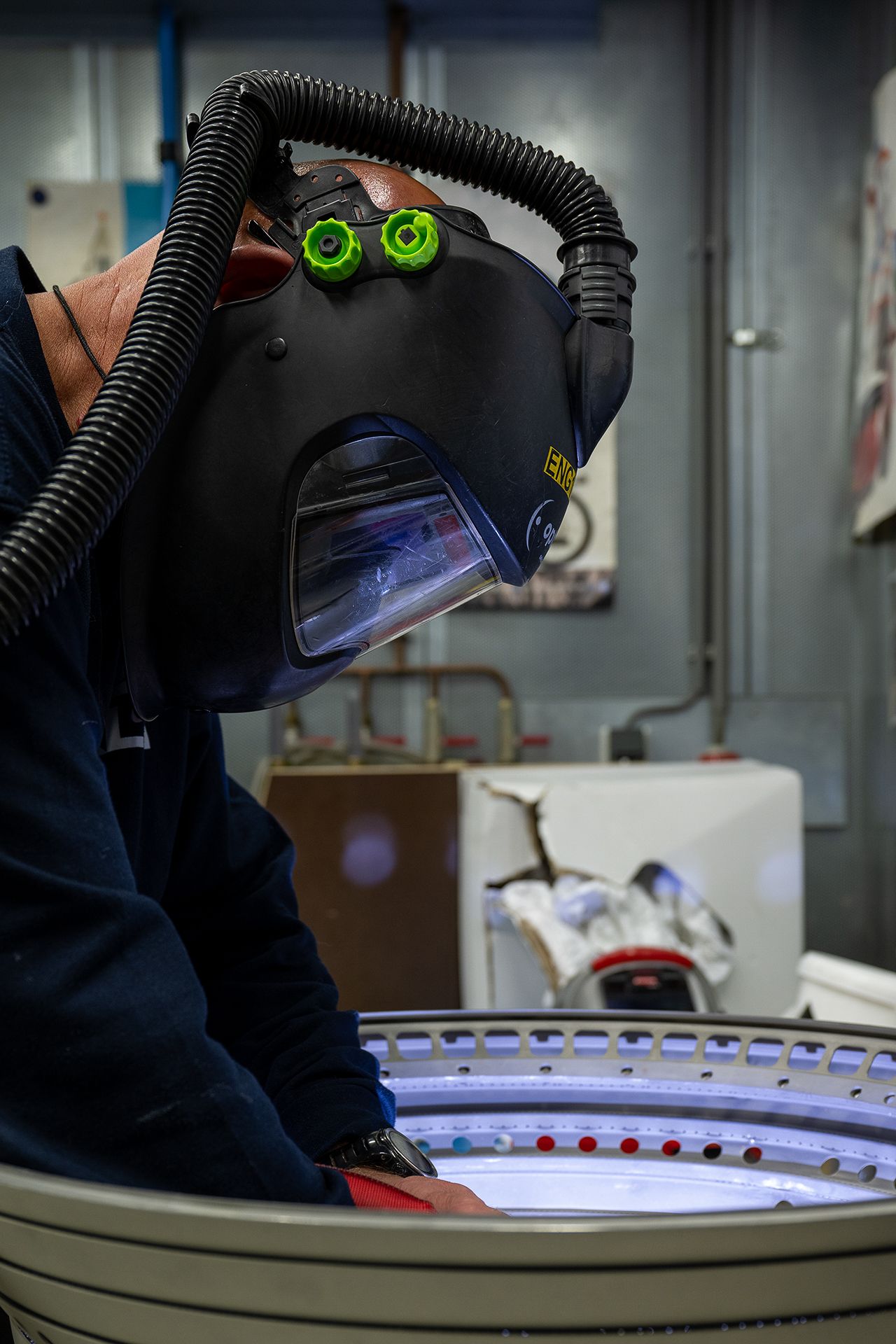 A person wearing protective gear and a full-face mask is working on a large round metal component.
