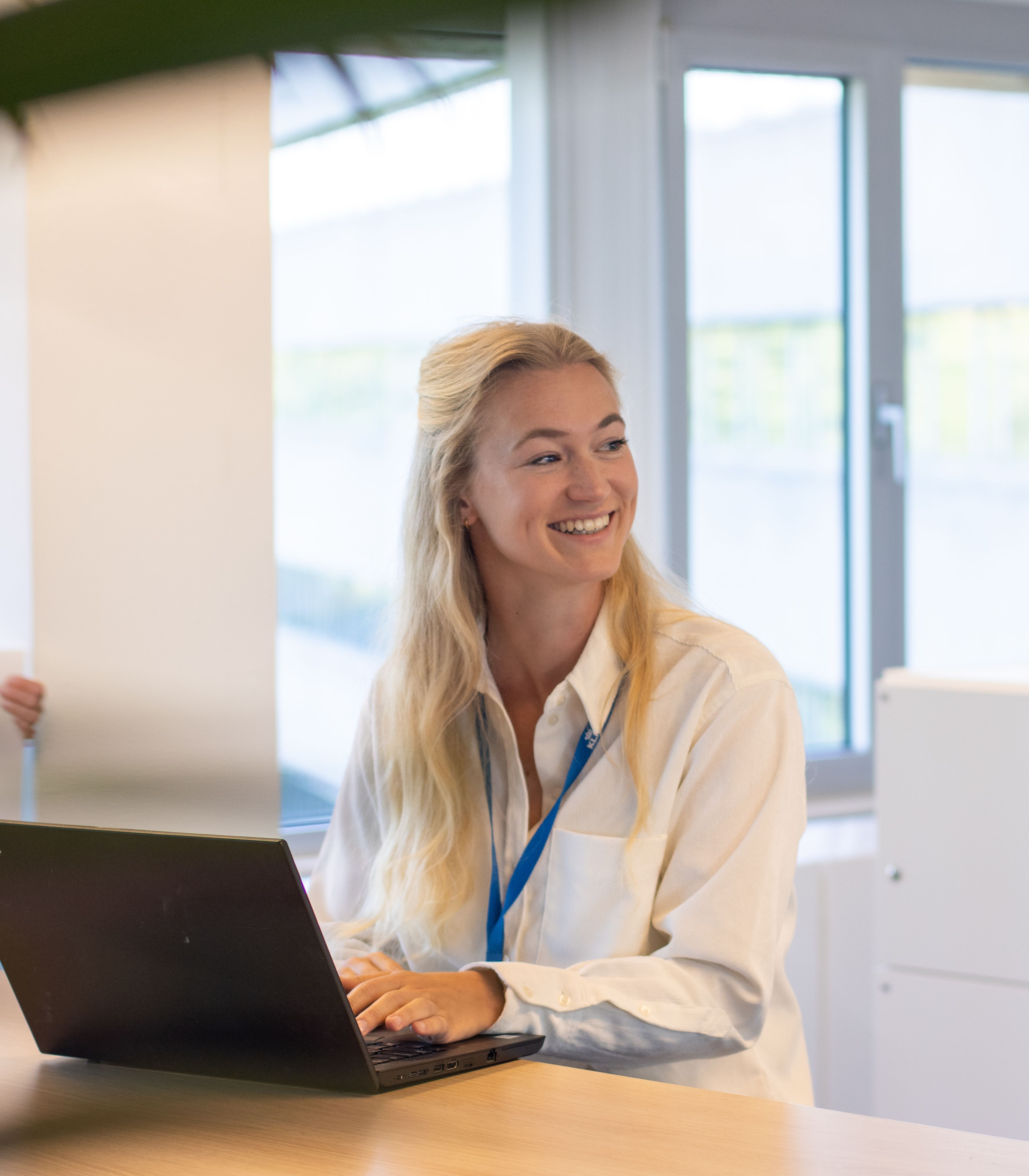 Vrouw lachend aan het werk met een laptop in een lichte kantoorruimte, met een collega op de achtergrond die een grote papierrol vasthoud