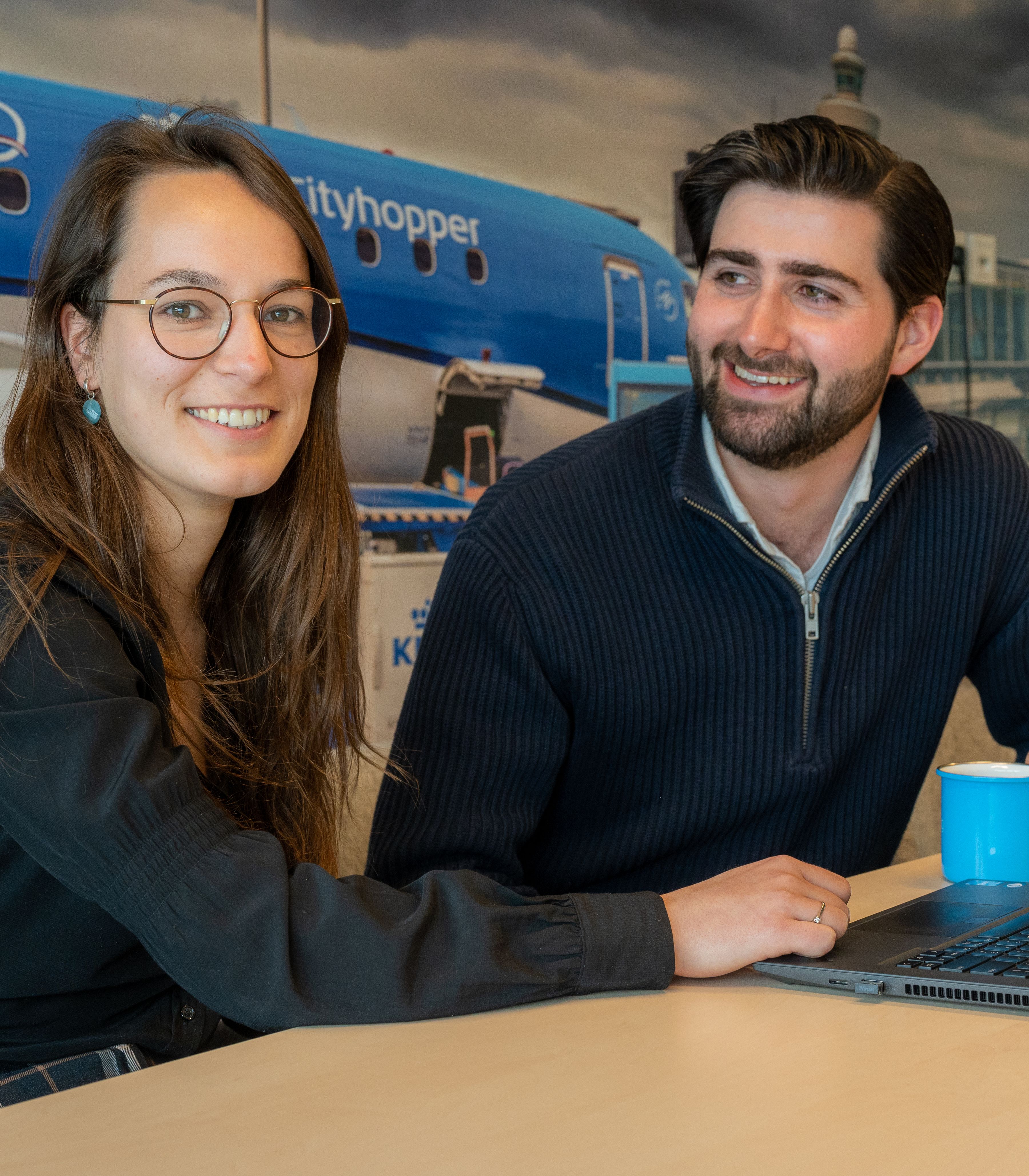 Twee collega’s aan tafel met laptop en koffiekopje, vliegtuigafbeelding op de achtergrond