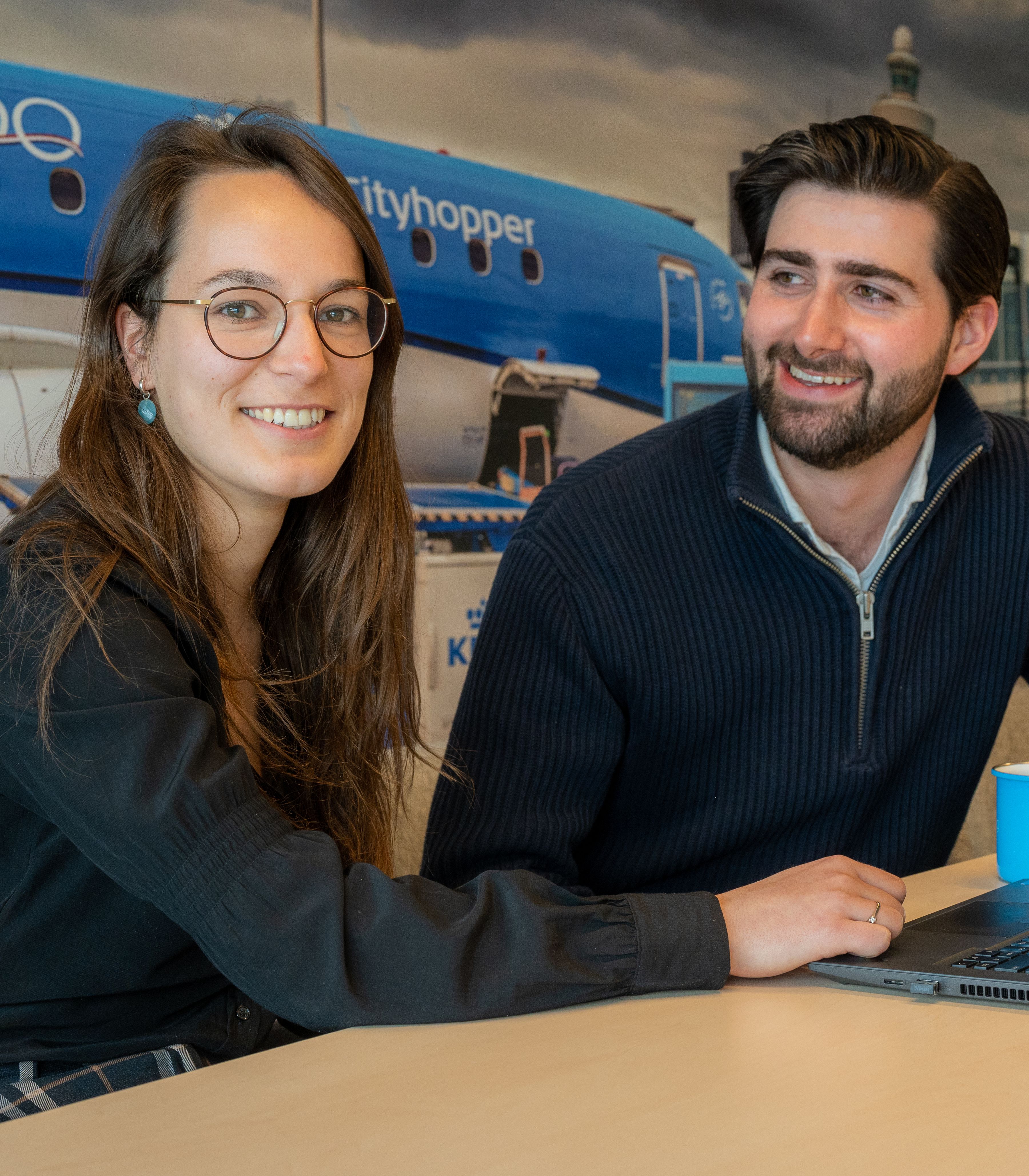 Two colleagues at a table with a laptop and coffee cup, aircraft image in the background.
