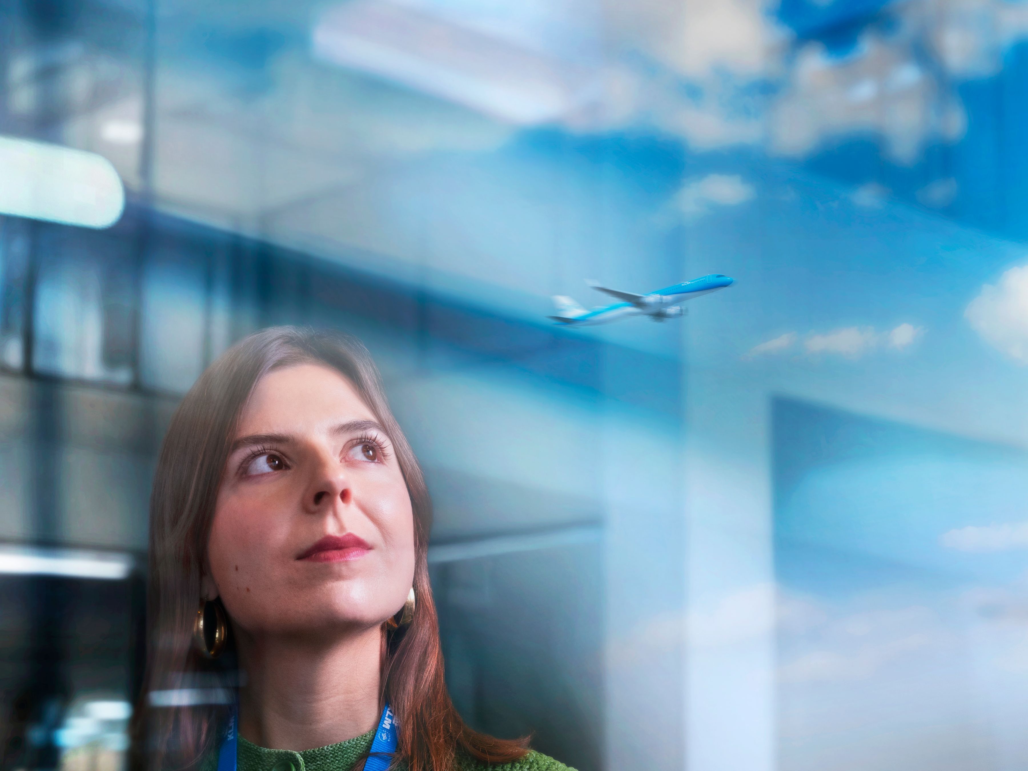 Een vrouwelijke KLM-collega kijkt uit een groot raam met een vertrekkend vliegtuig in de reflectie.