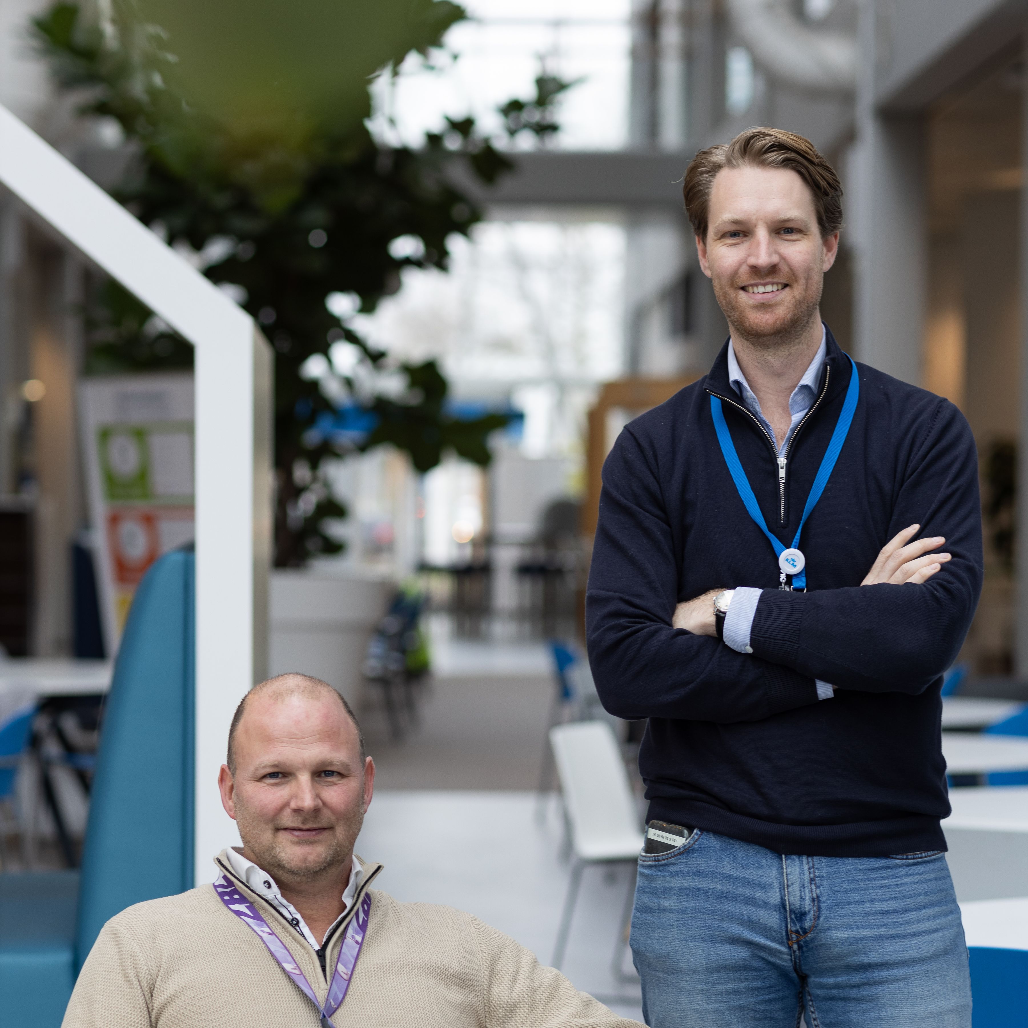 Twee mannen met een keycord, één zittend in een stoel en één staand erachter, in een moderne binnenruimte.