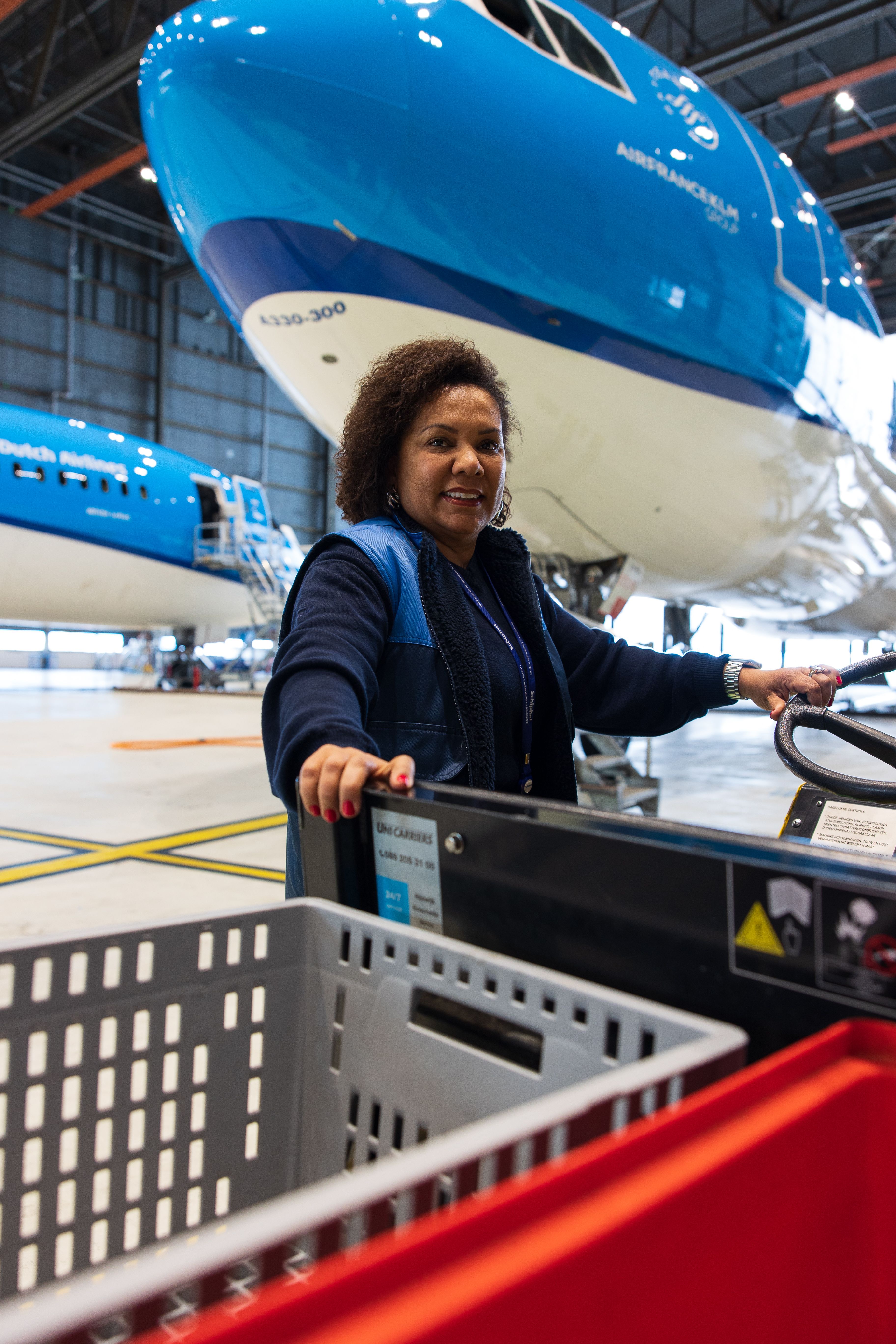 A woman in a hangar is driving a transport cart, with a KLM aircraft in the background.