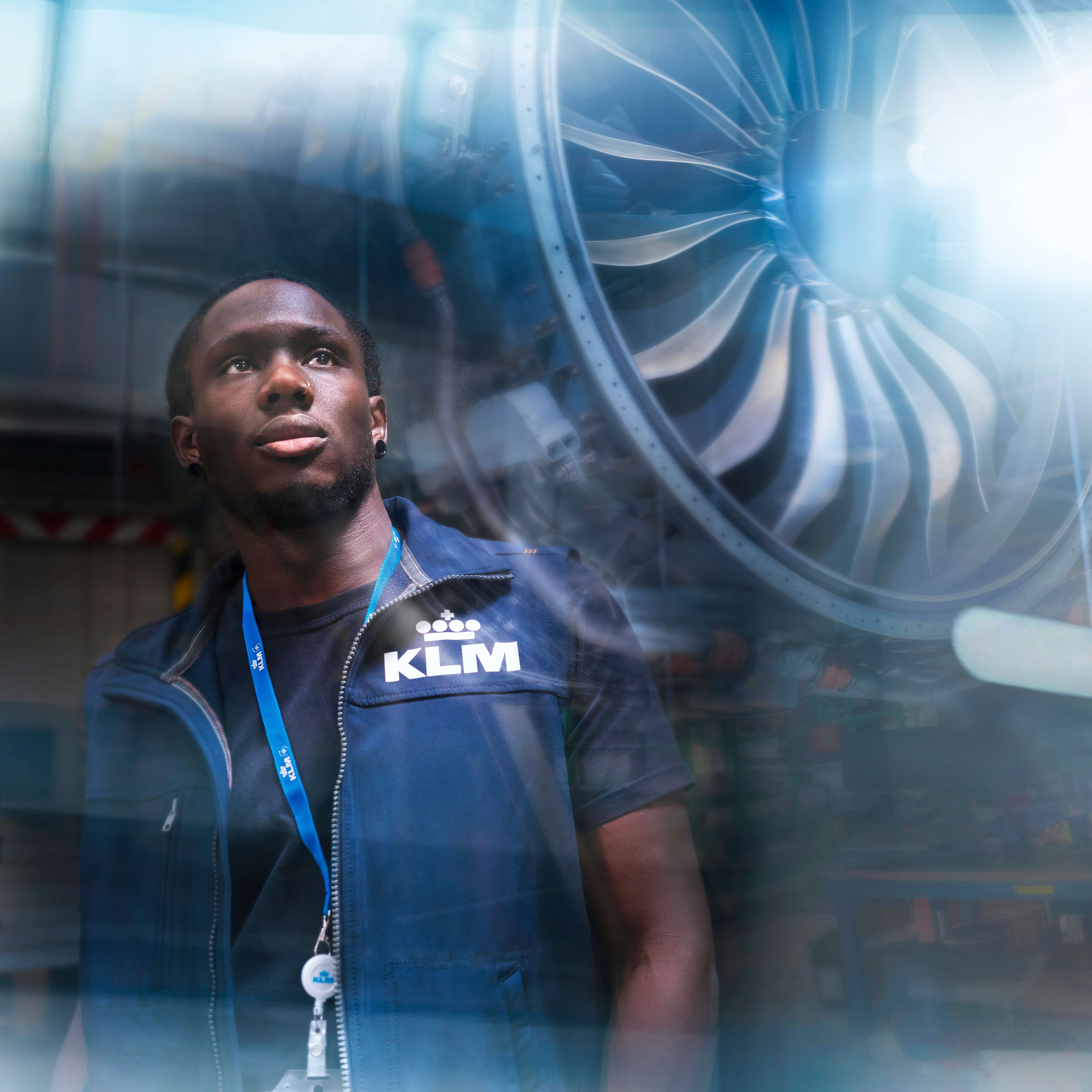 Man wearing a KLM vest and lanyard standing in front of an aircraft engine, with reflections on the glass.