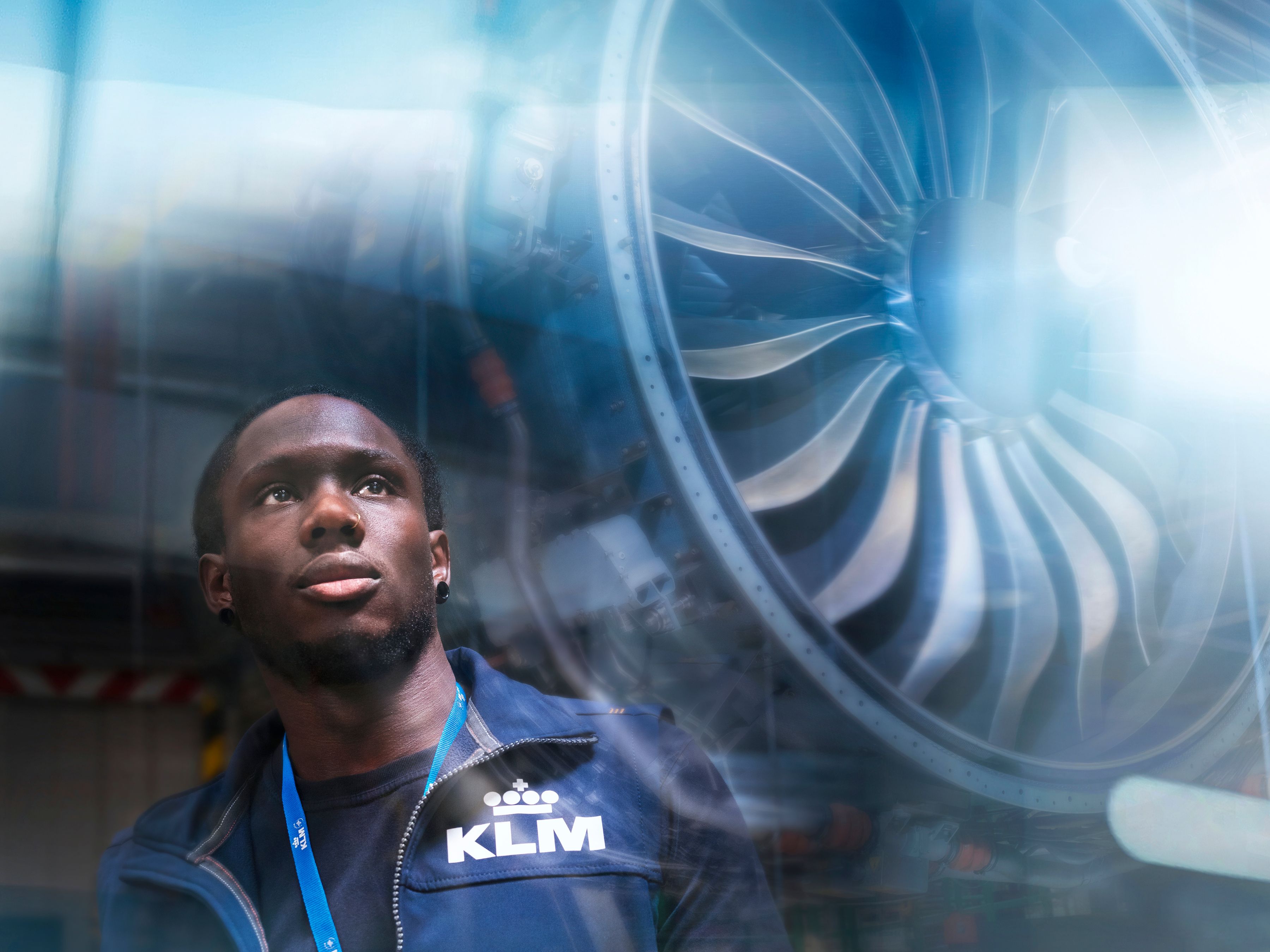 Man wearing a KLM vest and lanyard standing in front of an aircraft engine, with reflections on the glass.