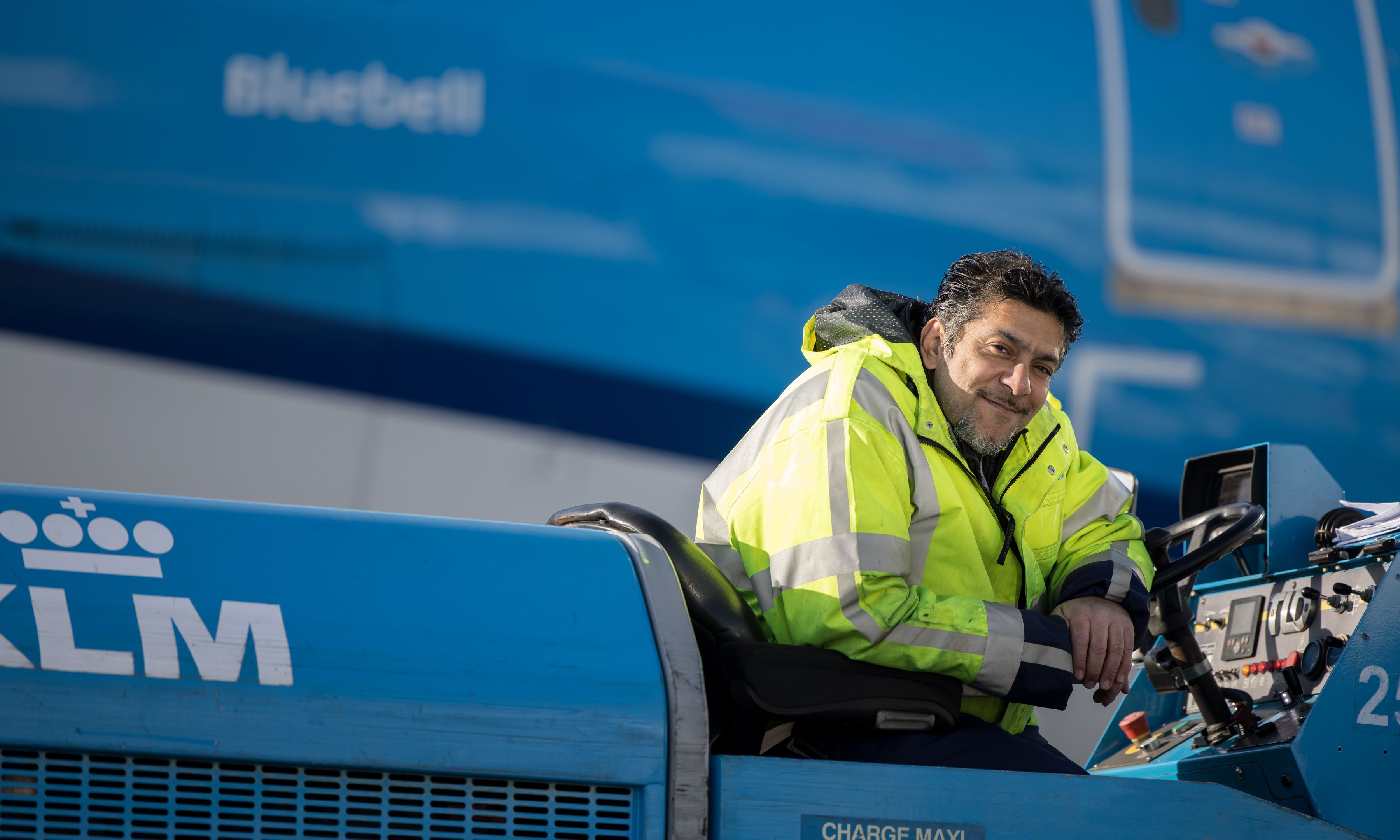 Ground staff member posing, he's sitting on his towtruck in front of a plane