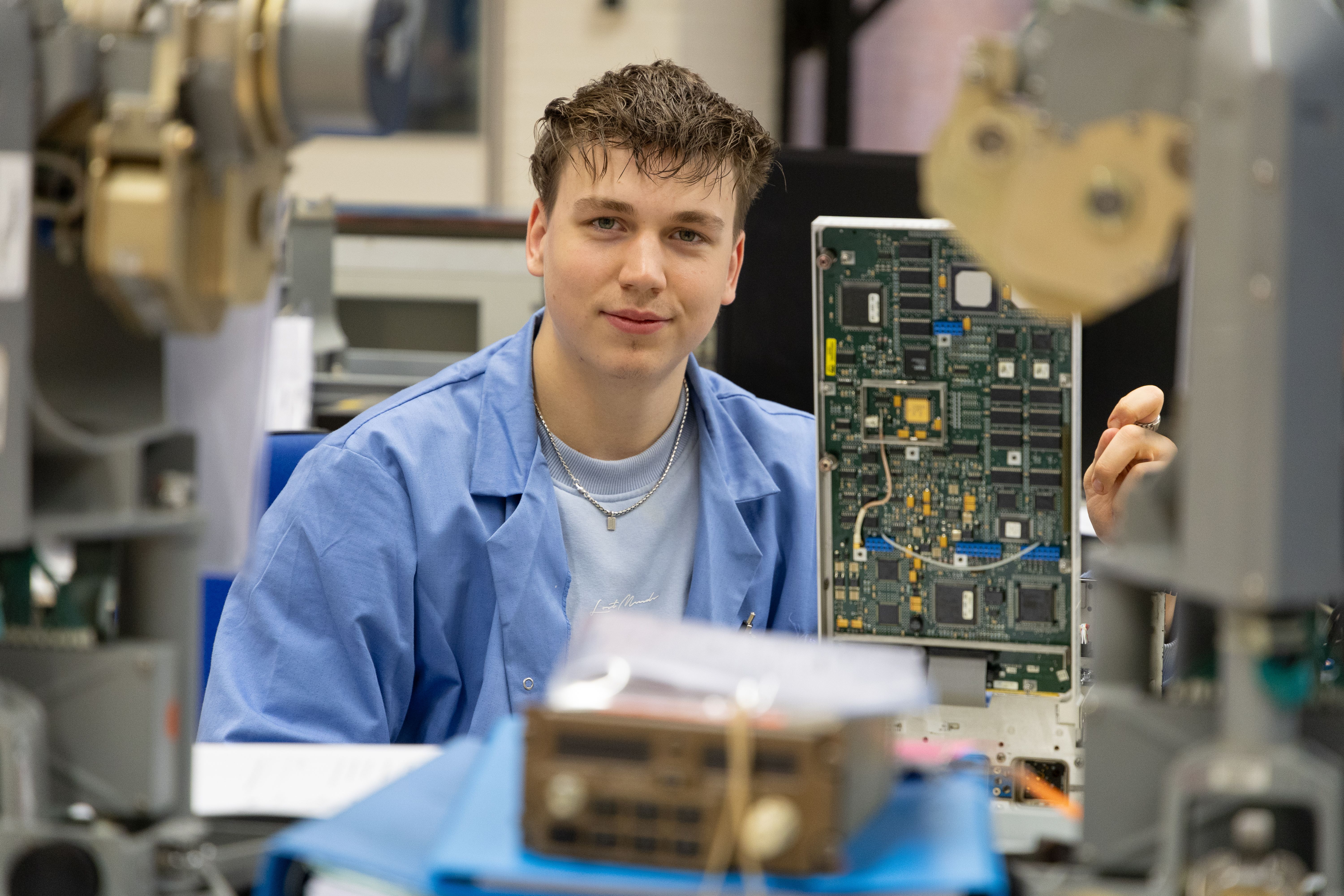 Young man in a blue lab coat holding up an electronic circuit board in a technical workspace