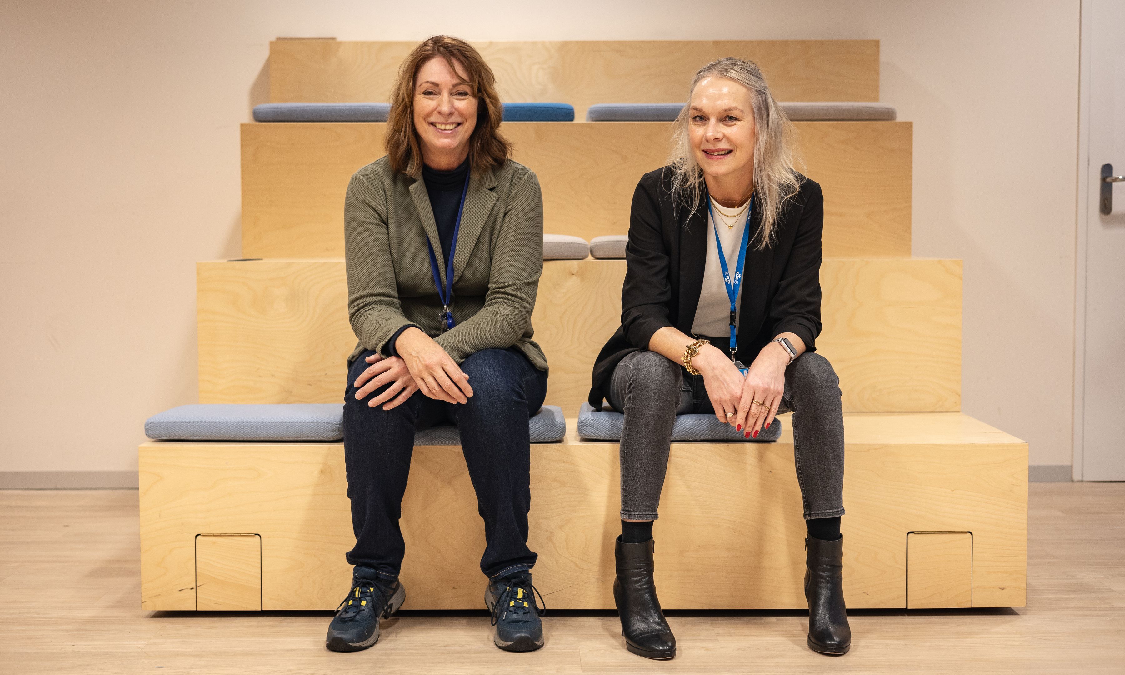 Two women with lanyards sitting on wooden steps with cushions, smiling at the camer
