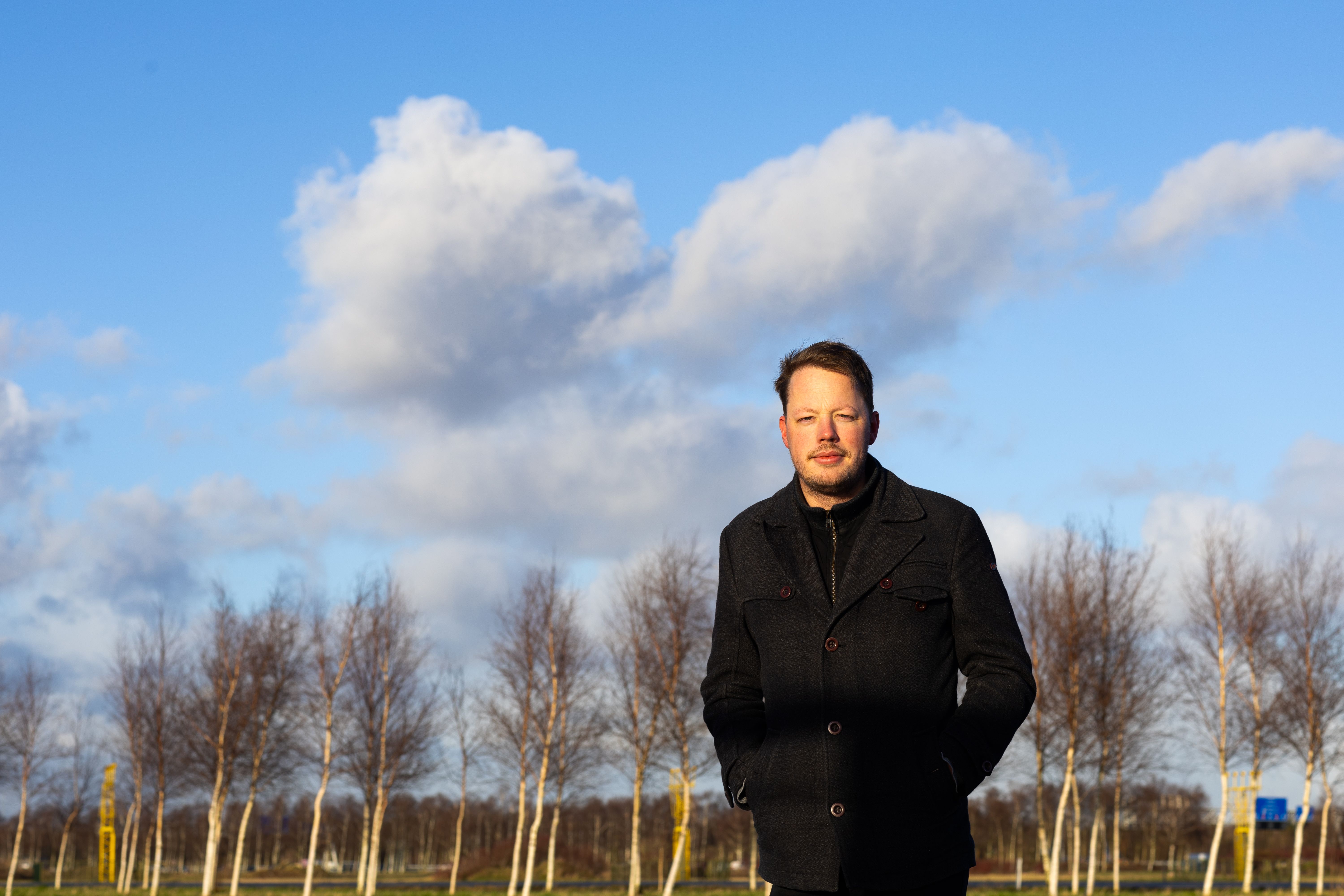 KLM-kantoor-collega staat voor een rij bomen met een blauwe lucht op de achtergrond.