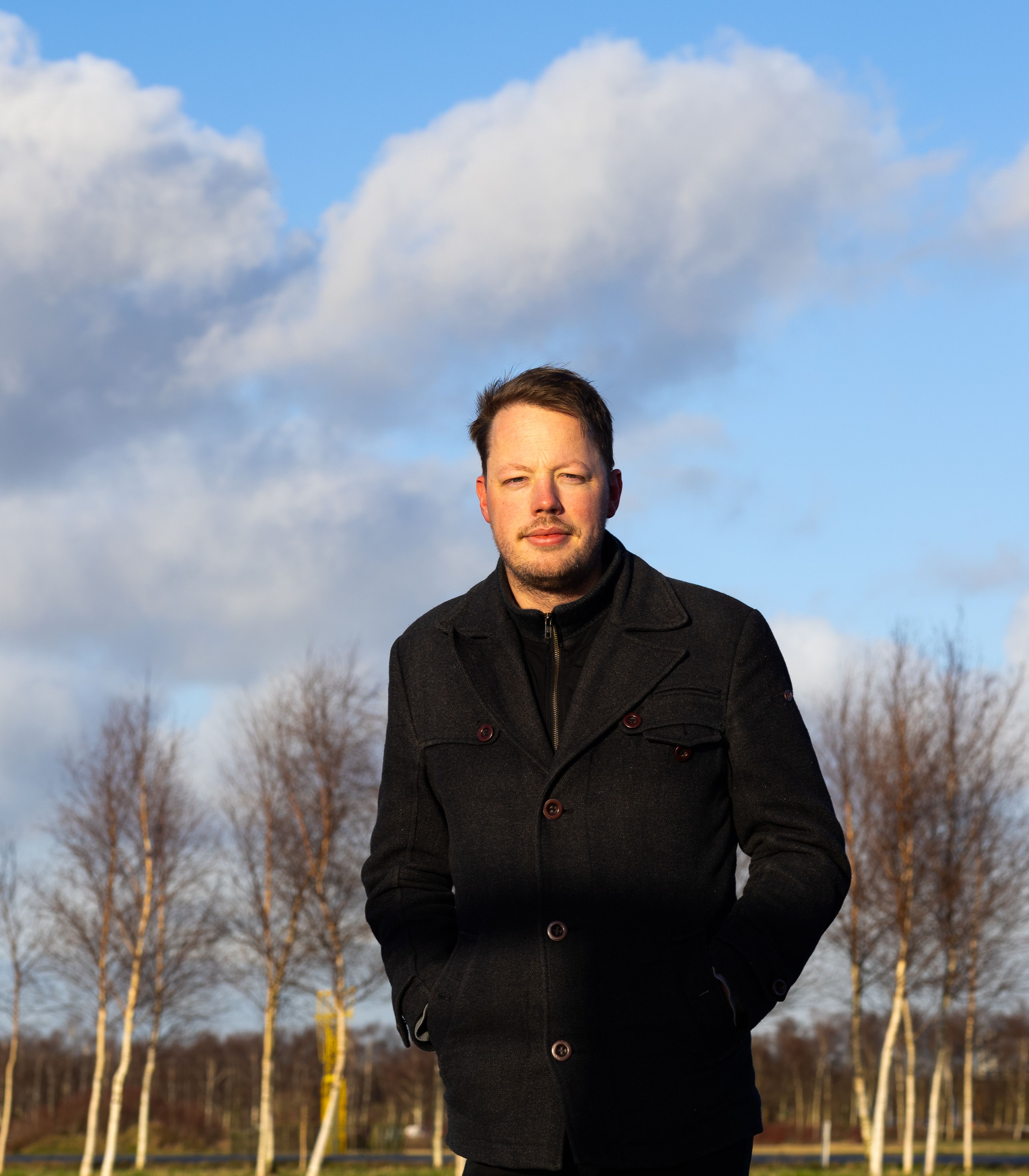 KLM-kantoor-collega staat voor een rij bomen met een blauwe lucht op de achtergrond.