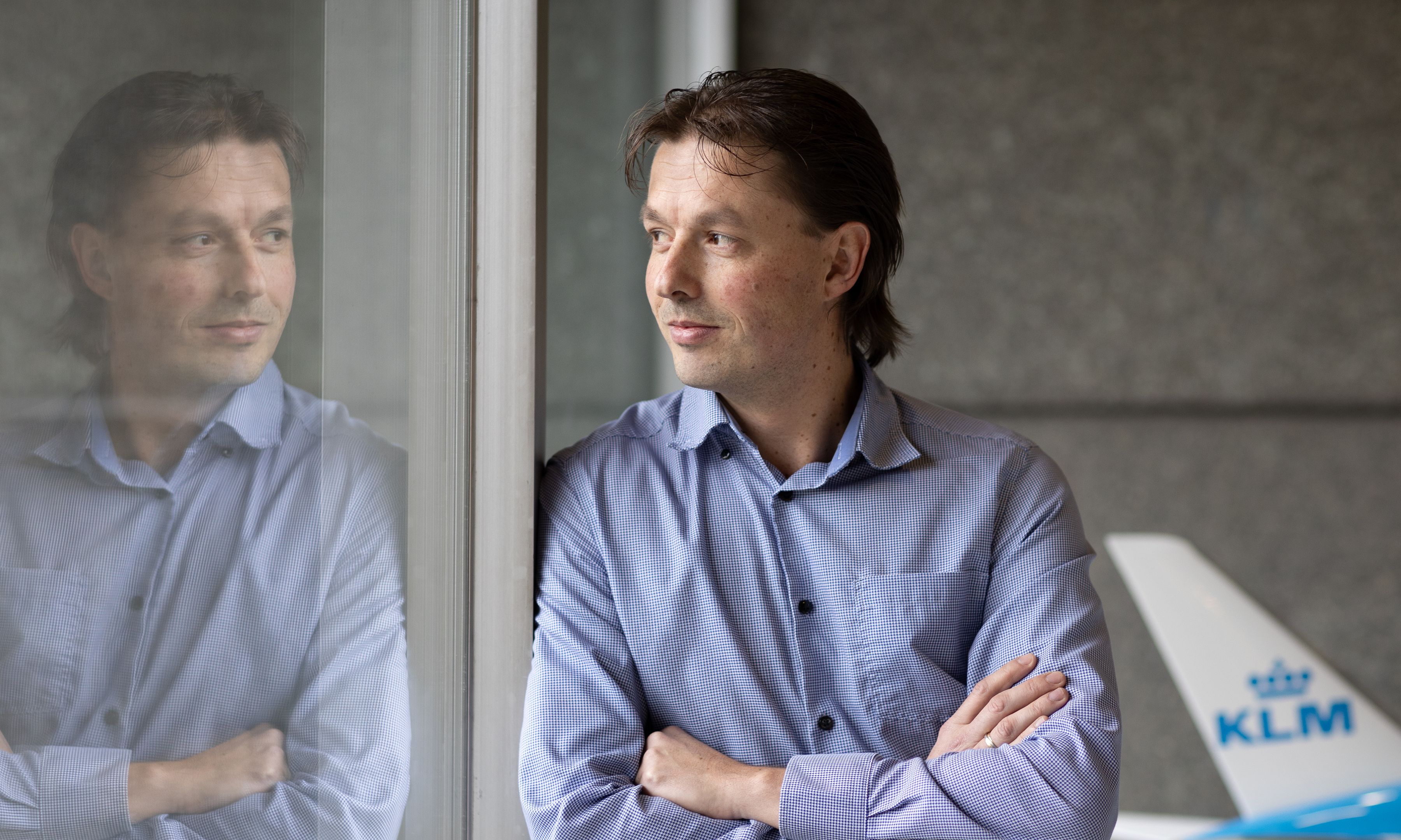 Man met gekruiste armen die uit een raam kijkt, met zijn spiegelbeeld zichtbaar en een modelvliegtuig op de achtergrond.