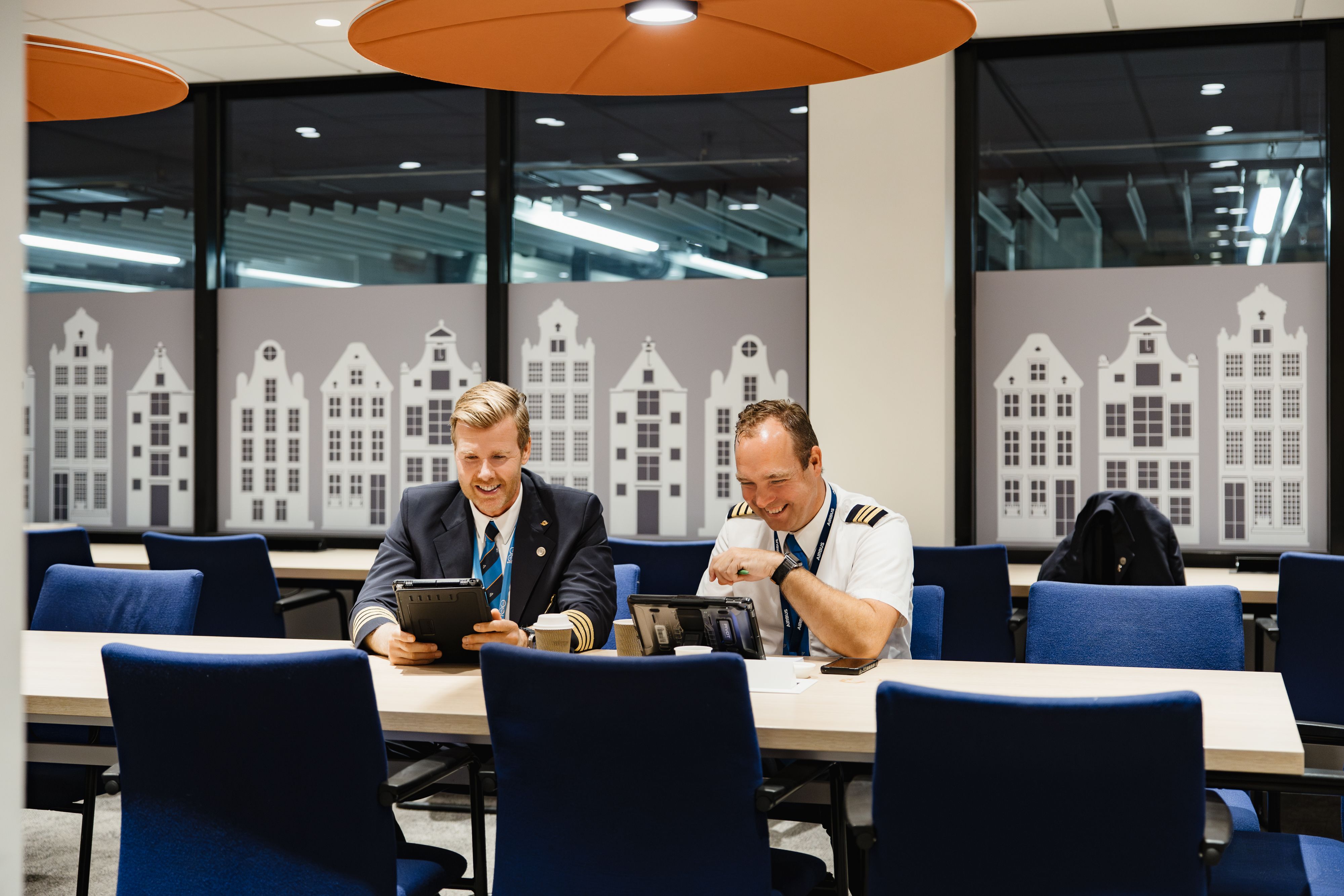 Twee piloten doen de vluchtvoorbeiding, een heeft een ipad in zijn hand de ander moet lachen. Ze zitten aan een lange tafel met KLM huisjes op de achtergrond.