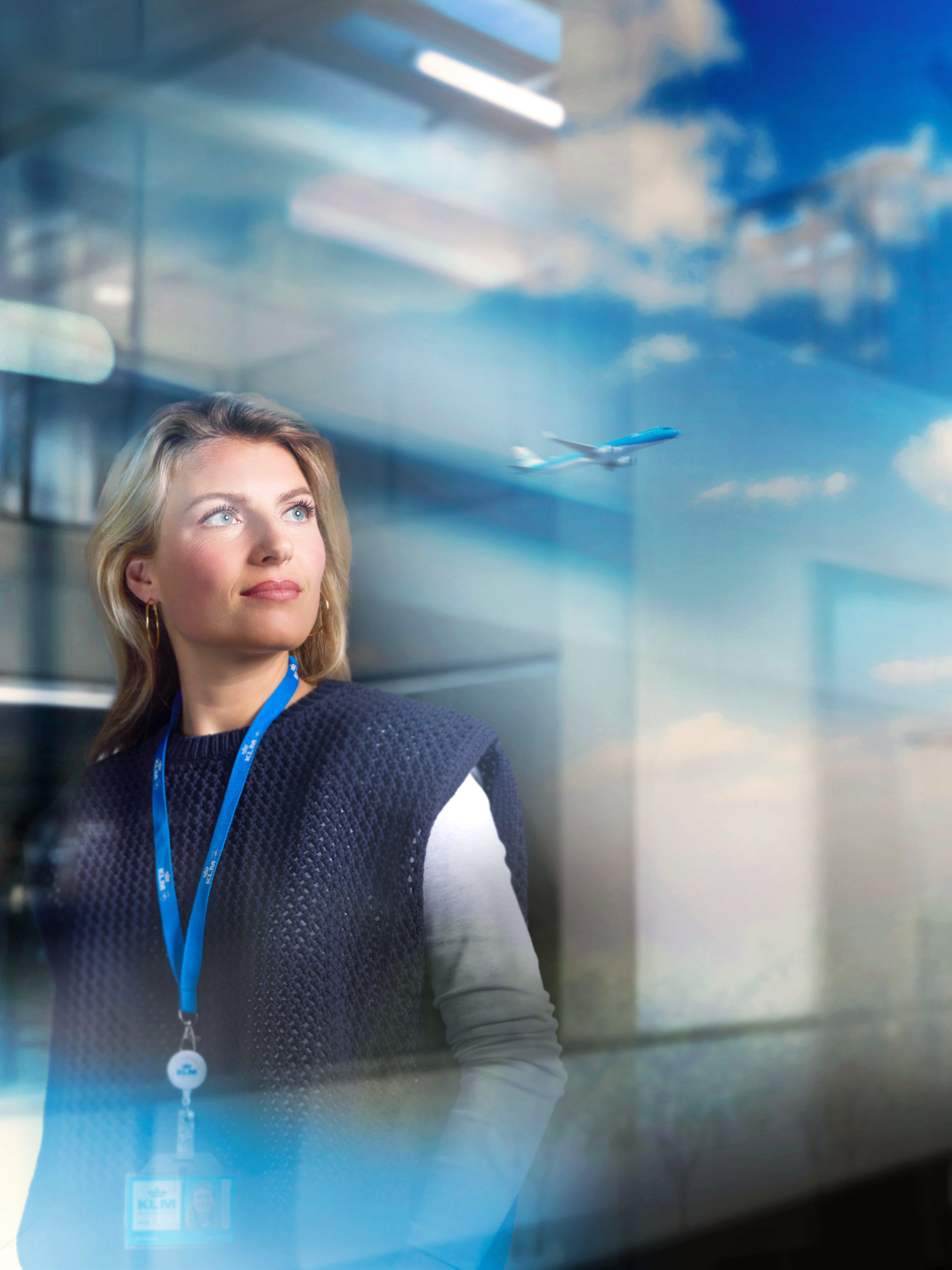 Een vrouwelijke KLM-collega kijkt uit een groot raam met een vertrekkend vliegtuig in de reflectie.