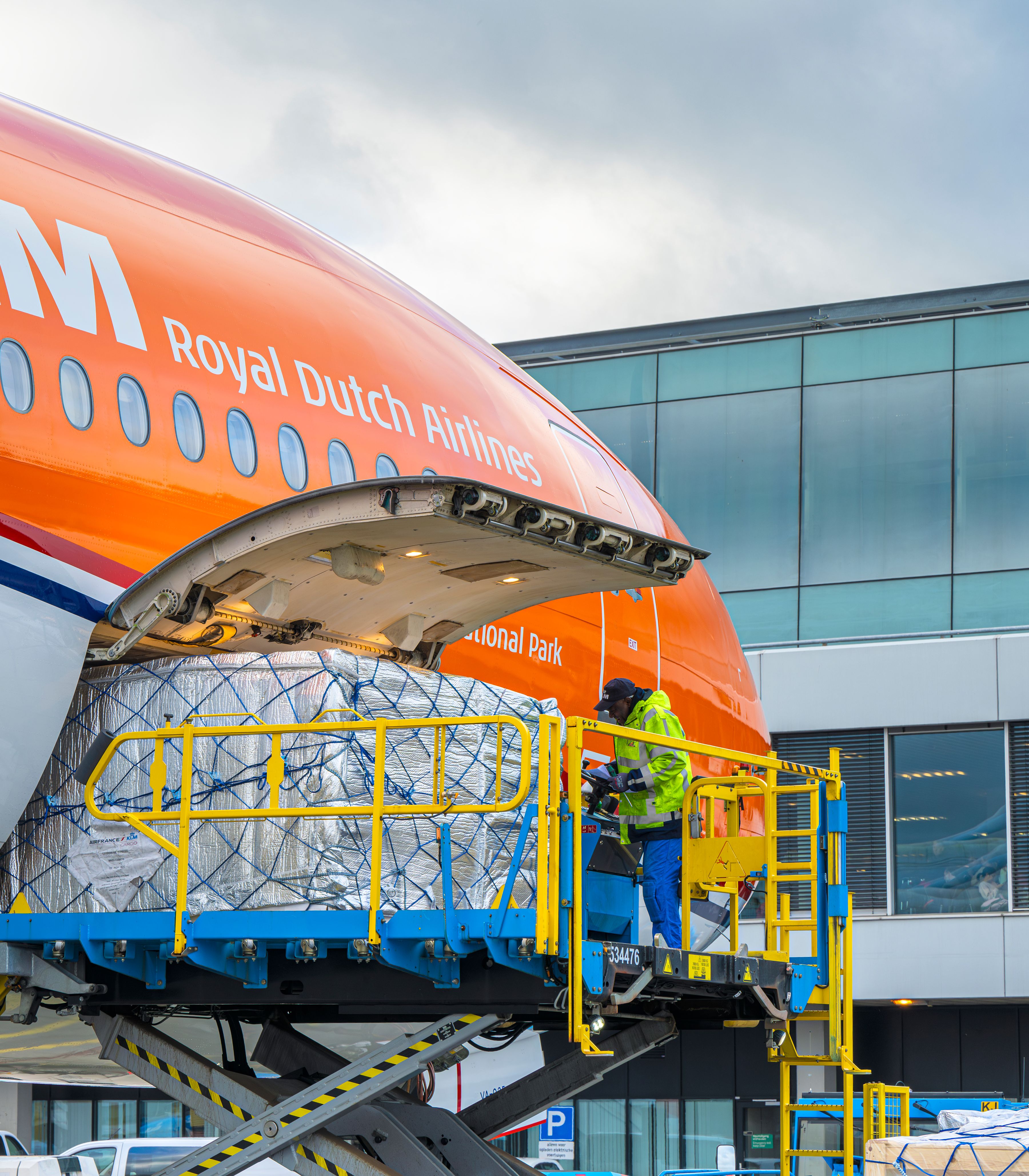 Vracht wordt via een schaarlift in de neus van een oranje KLM-vliegtuig geladen op de luchthaven.