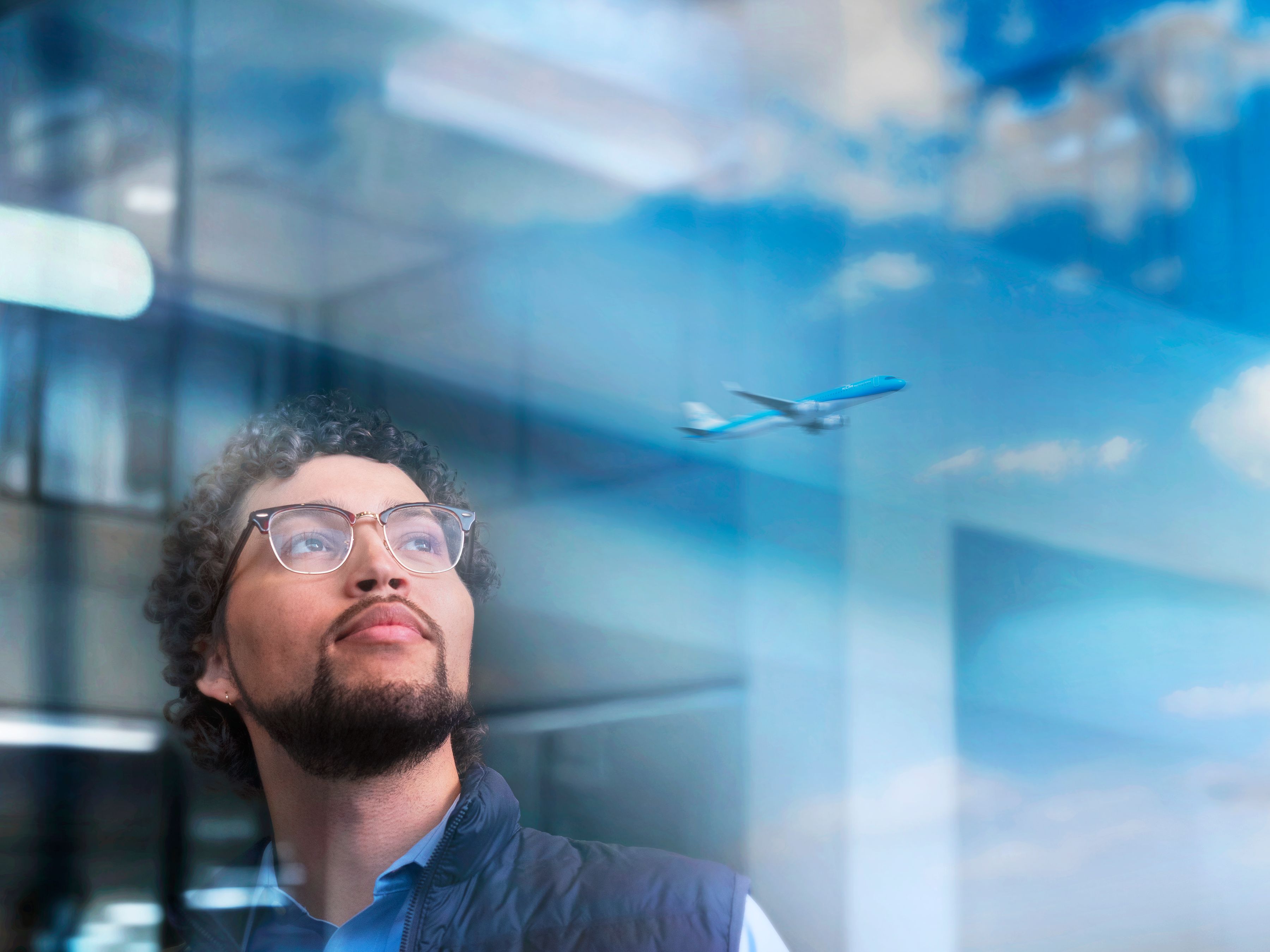 Man with glasses and a lanyard looking up, with reflections of clouds and an airplane in the window.