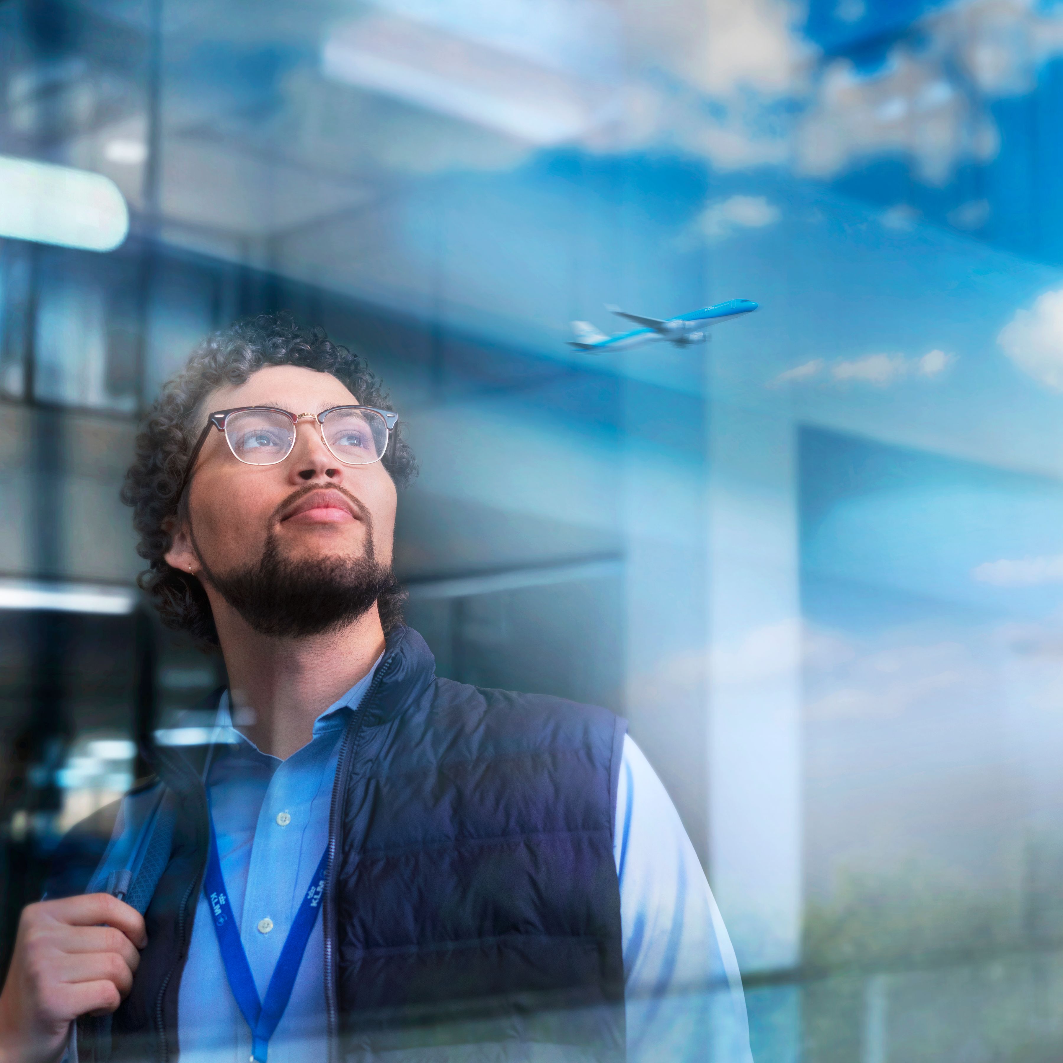 Man with glasses and a lanyard looking up, with reflections of clouds and an airplane in the window