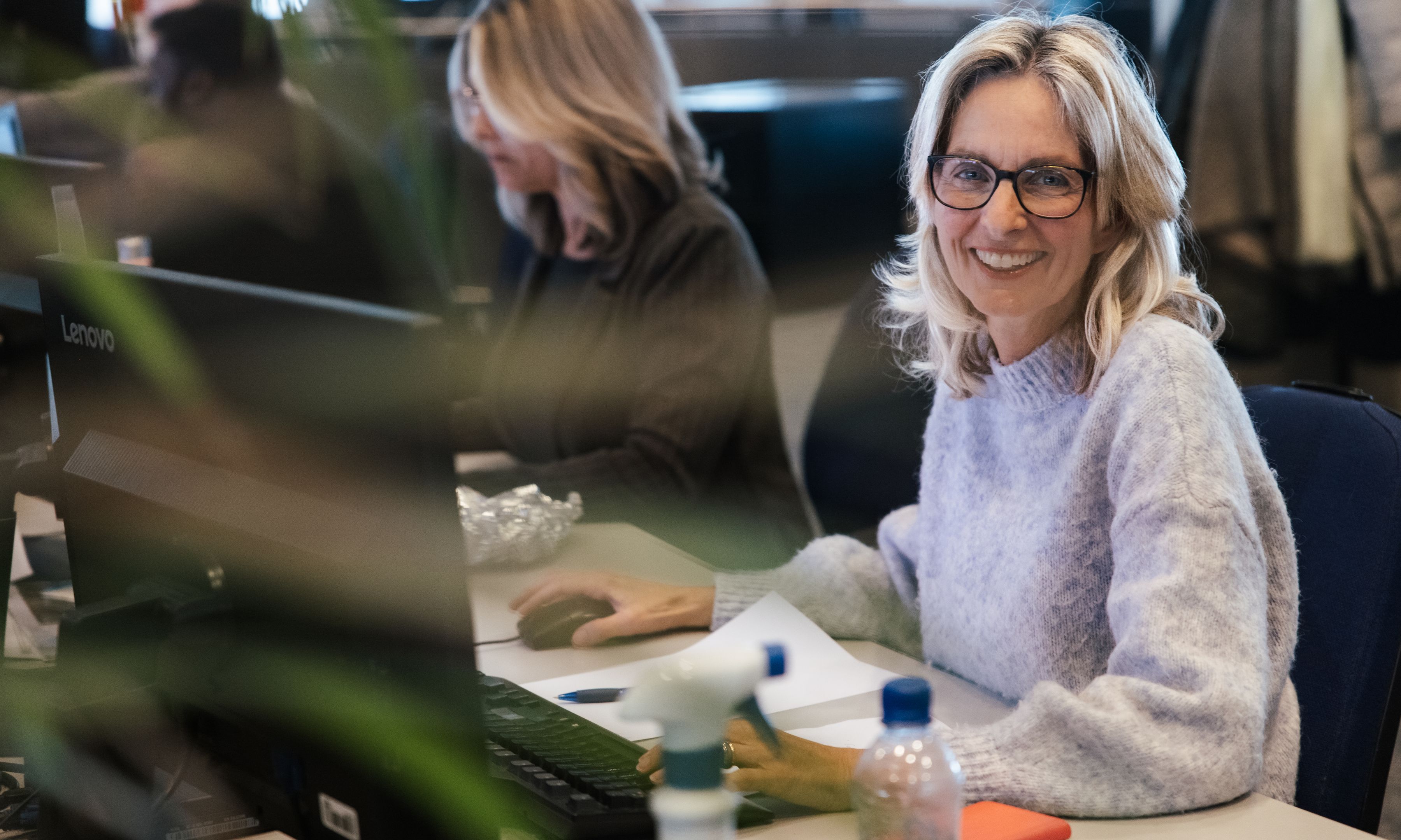 Een lachende vrouw met een bril en een lichte trui zit aan een bureau op kantoor, met op de achtergrond een andere persoon die aan het werk is.