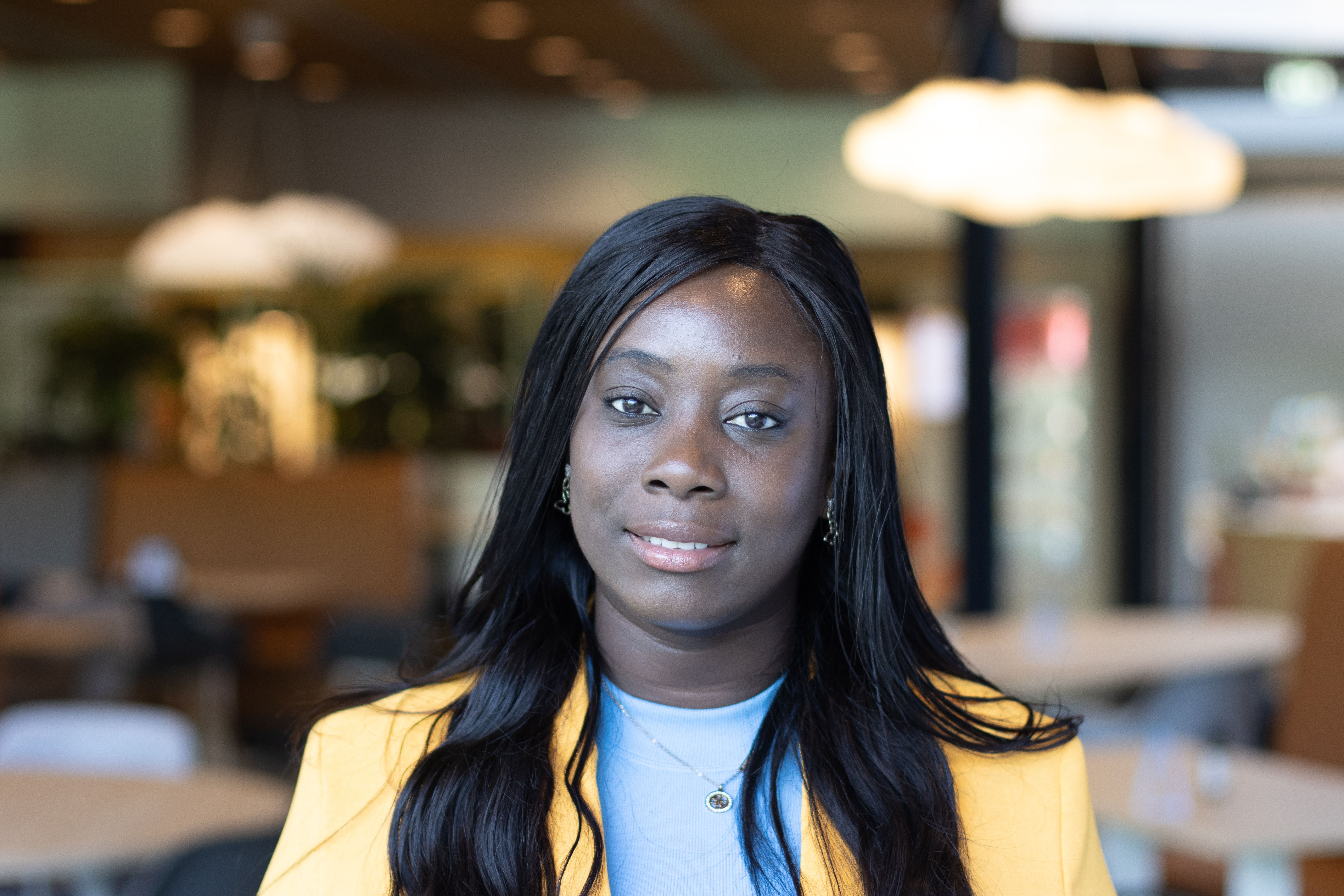Vrouw met lang donker haar in een gele blazer, poserend binnen met een vage achtergrond.