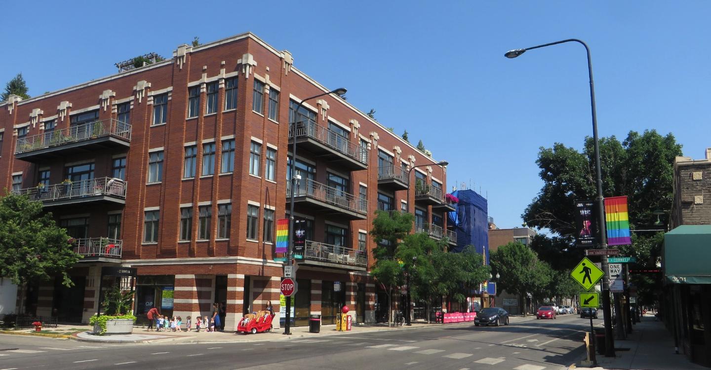 A Northalsted street corner with a brick apartment block, rainbow flags, and a clear summer sky