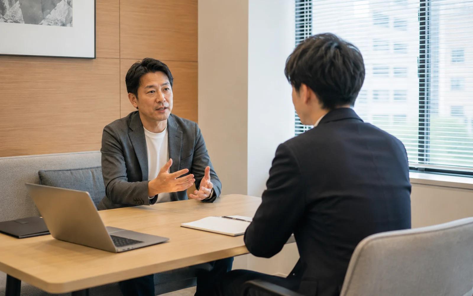 会議室のテーブルを挟み、ジャケット姿の男性が身ぶりを交えて話し、向かい側の社員が背を向けて聞いている場面